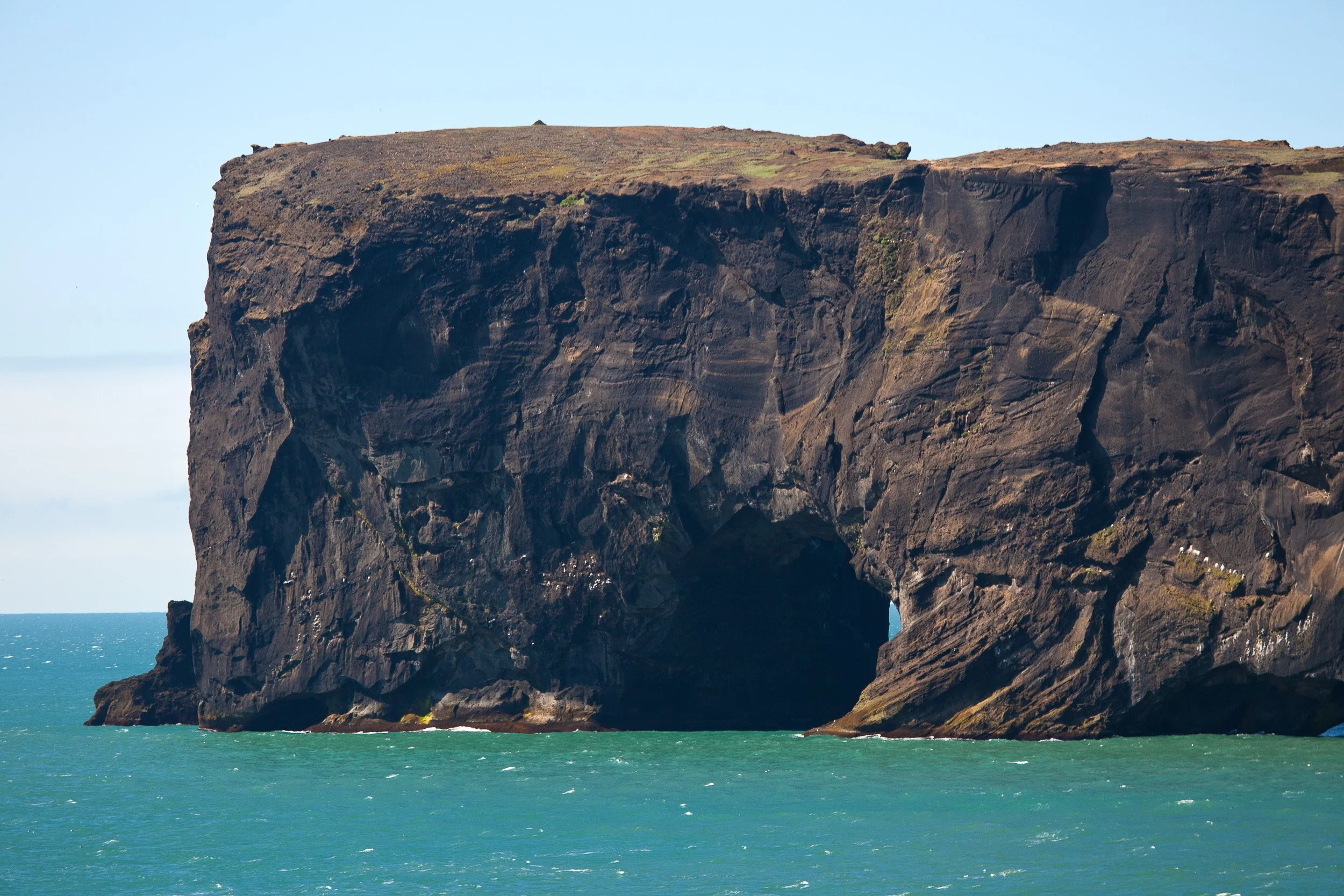 A hulking sea arch cut into dark volcanic cliffs, with turquoise Atlantic water surging below in bright summer light.