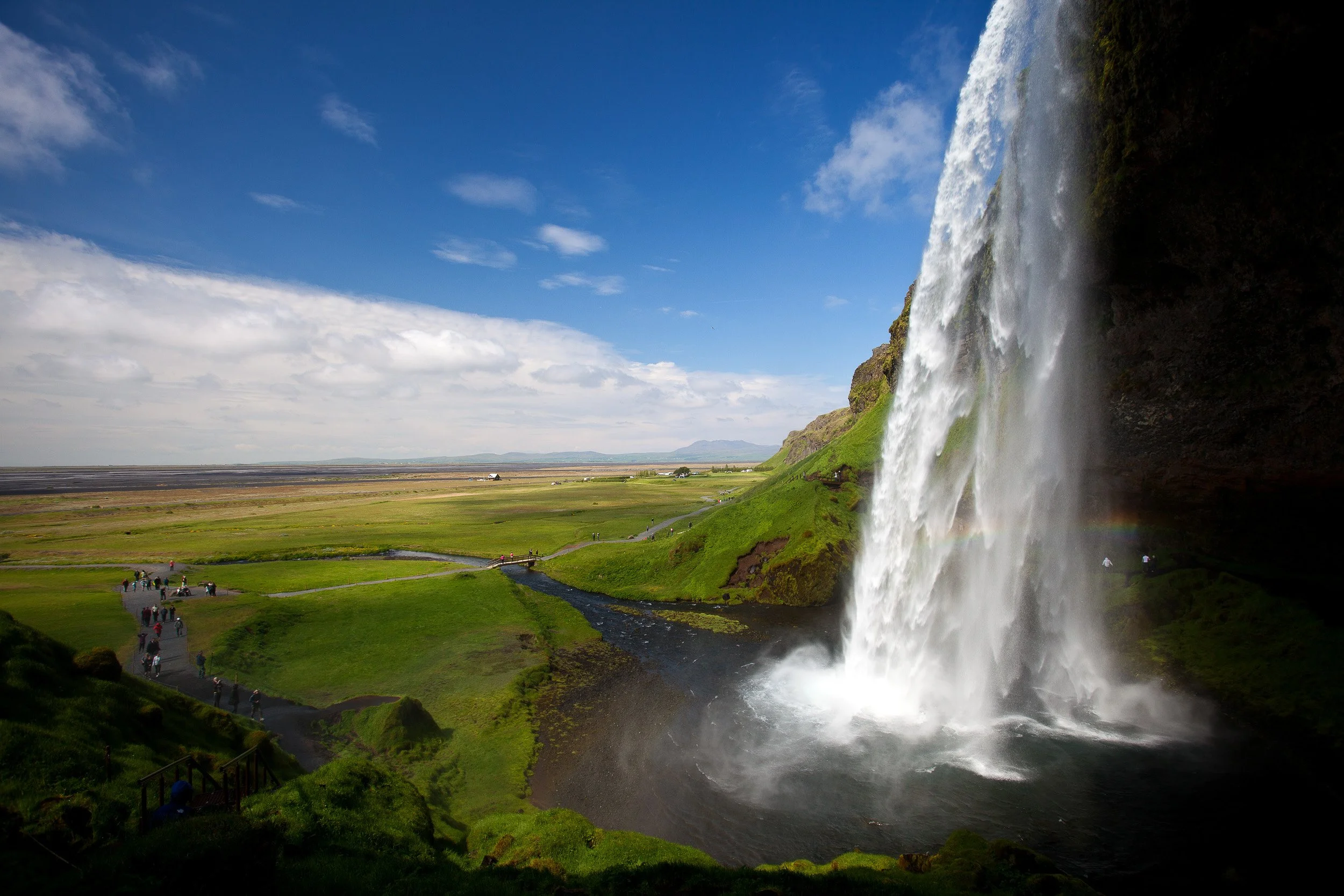Seljalandsfoss — the classic side view across the plains, with spray, sunlight and a faint rainbow hinting at Iceland’s sudden weather shifts.