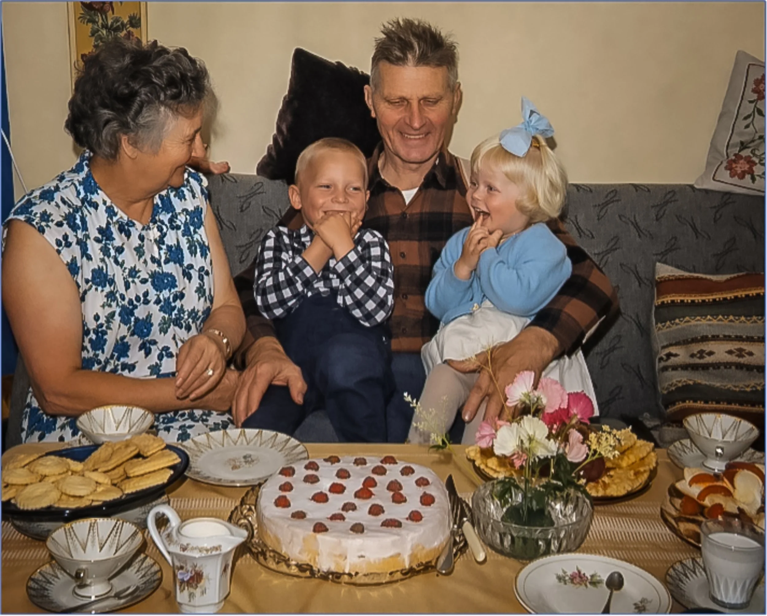 Erna og Ivar O. Husevåg sammen med barnebarna Ivar Husevåg og søsteren Elisabeth, trolig omkring 1966. Familien sitter rundt et festdekket bord med kake og kaffekopper under et besøk hos besteforeldrene i Husevåg.