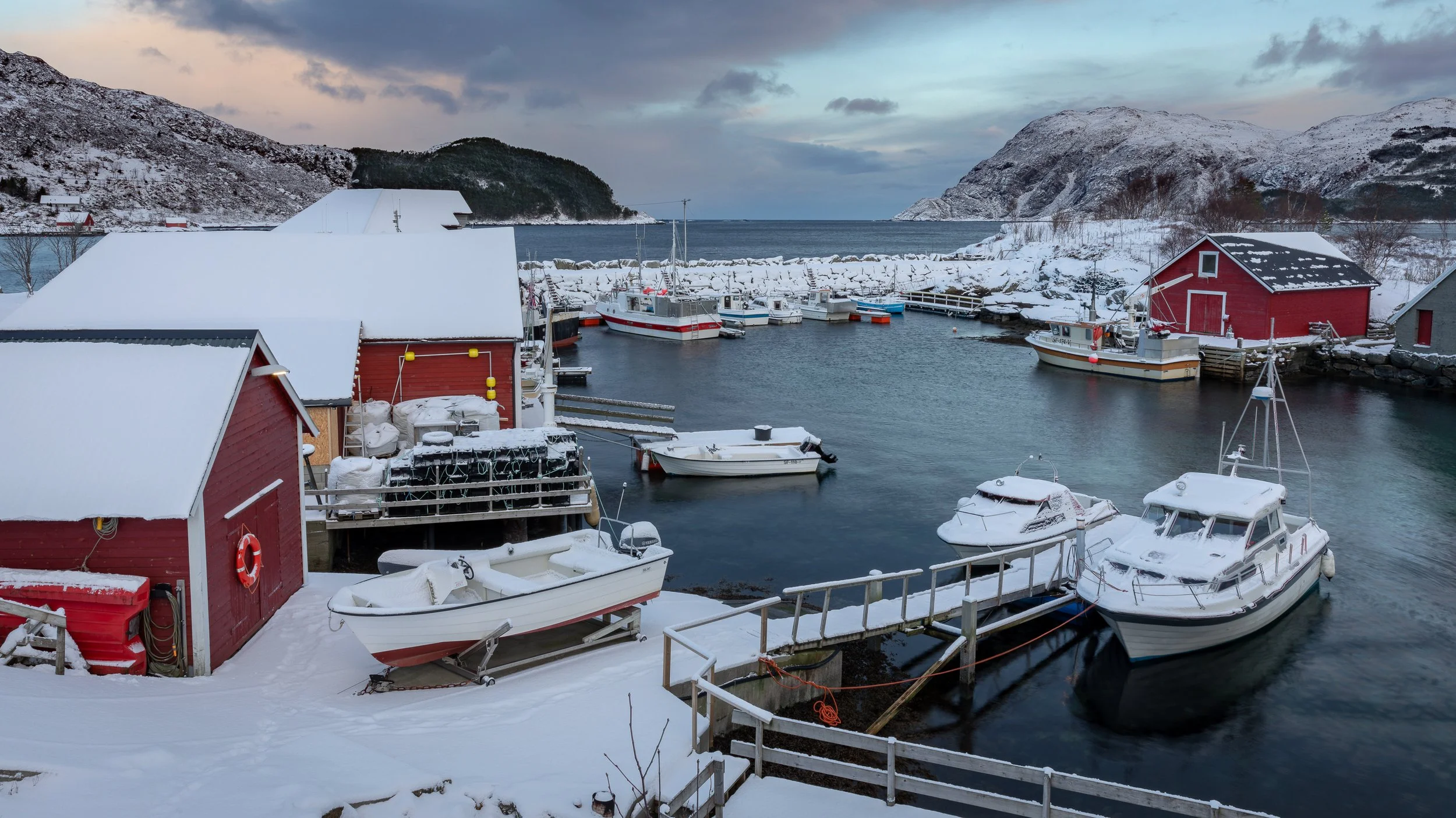 Vinterbilde av Vågen i Husevåg på Husevågøy, med røde sjøhus, småbåter, kaier, snødekte fjellsider og moloen som beskytter havna mot havet utenfor.
