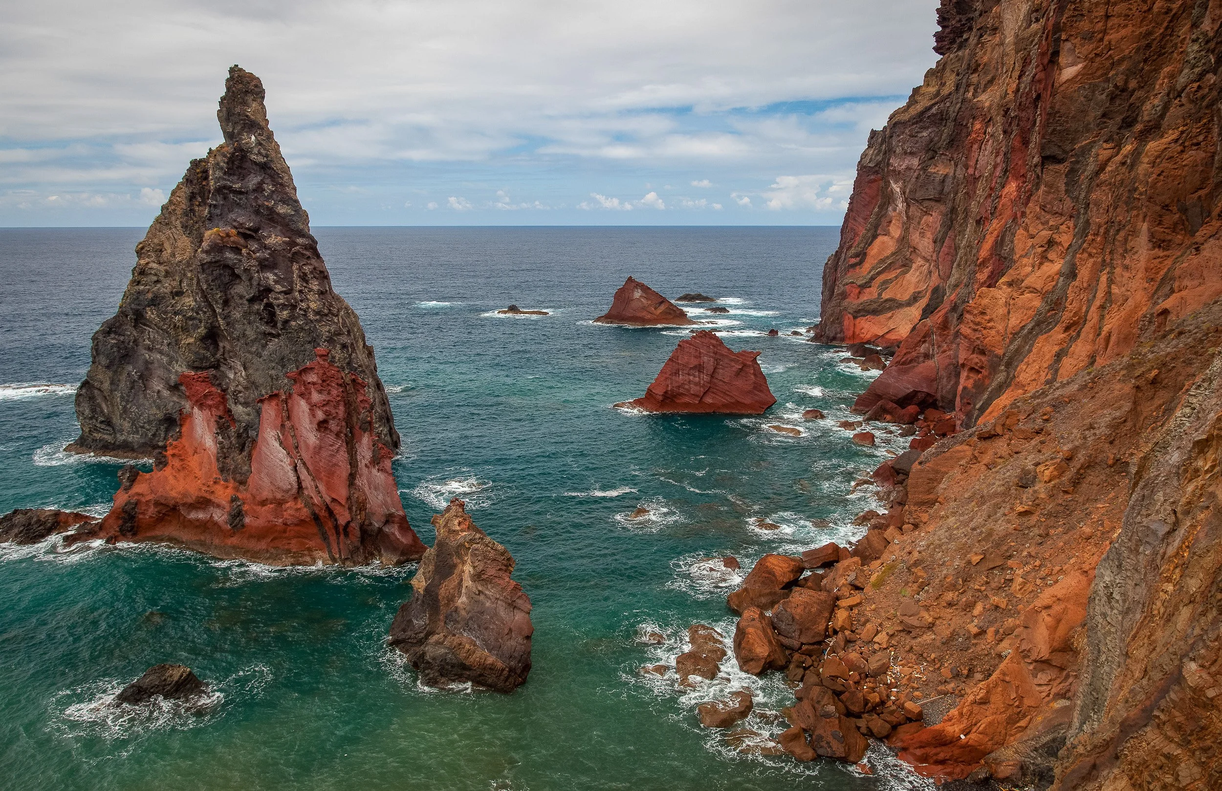 Dark sea stacks and red volcanic cliffs at Ponta de São Lourenço, with waves breaking into turquoise water below the exposed eastern headland.