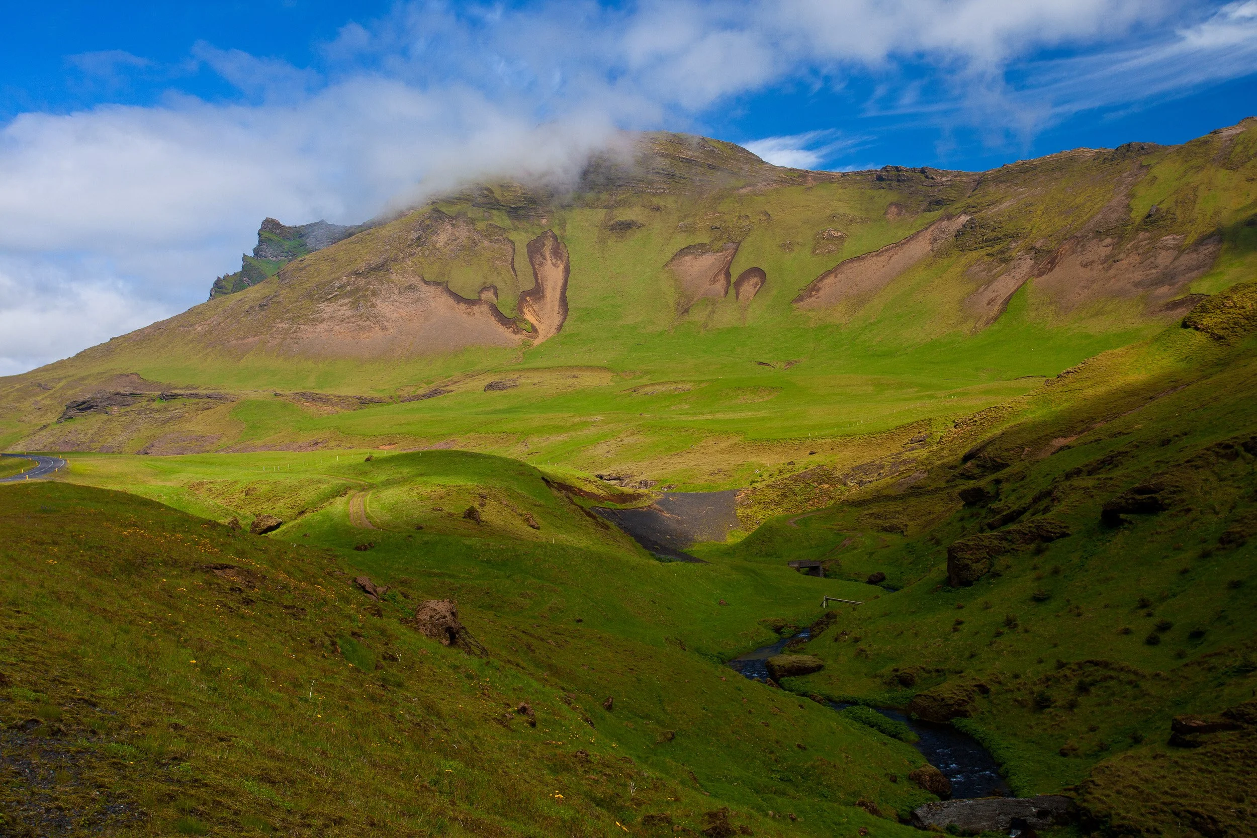 Velvet-green volcanic hills under fast-moving cloud, with a narrow stream cutting a dark line through the valley floor.