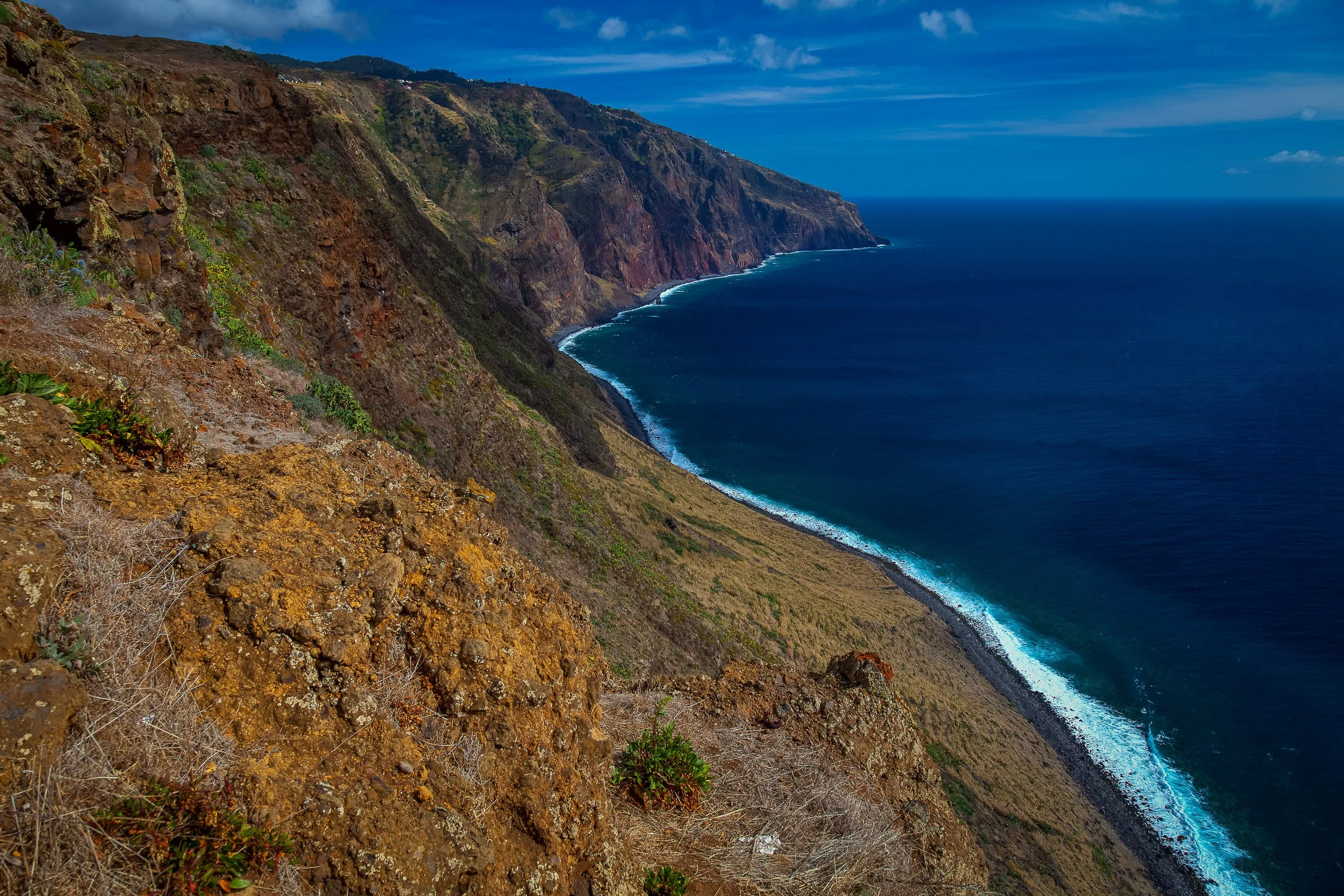 Steep coastal cliffs near Ponta do Pargo, where the Atlantic follows the shoreline in a pale ribbon of surf below dry slopes and scattered cloud.