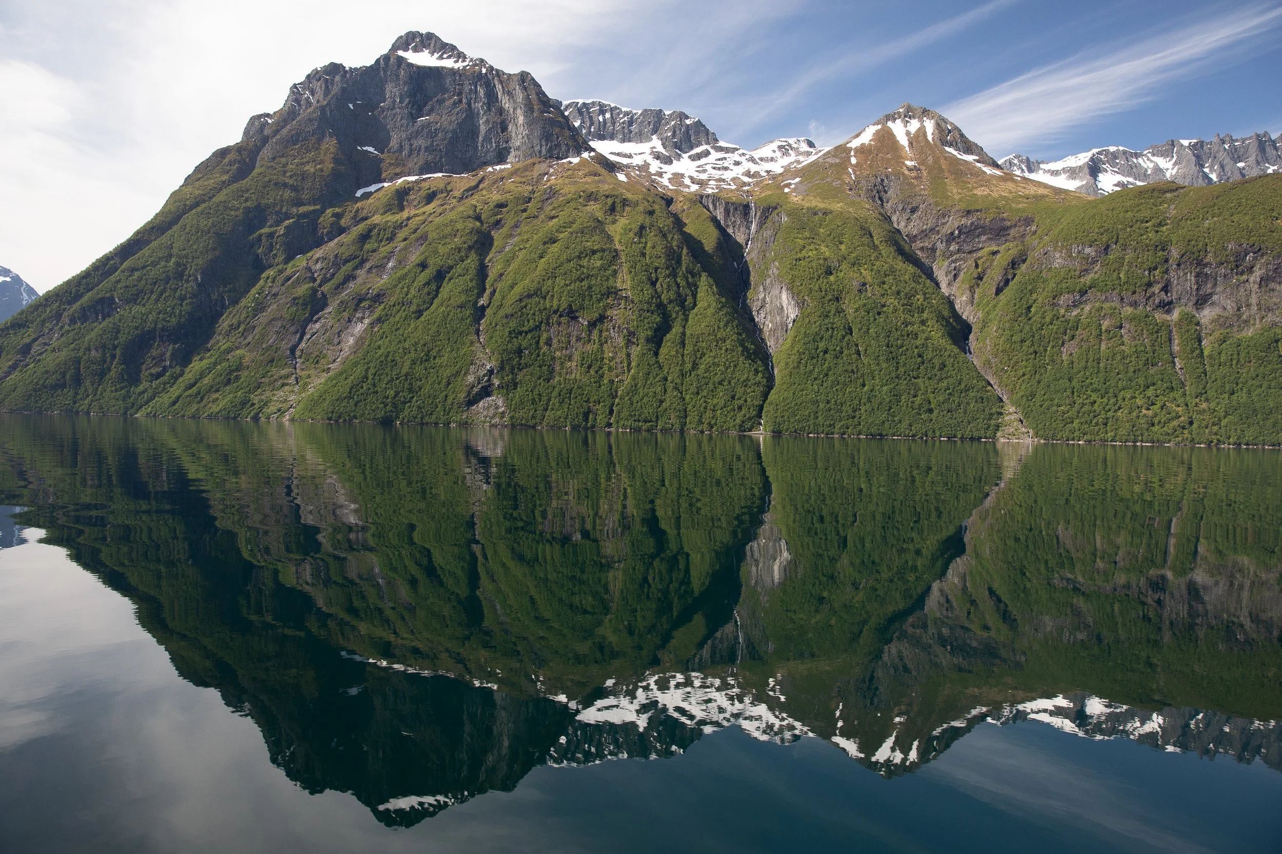Hjørundfjorden in stillness — mountain slopes mirrored in calm water, with small human details dwarfed by the landscape.