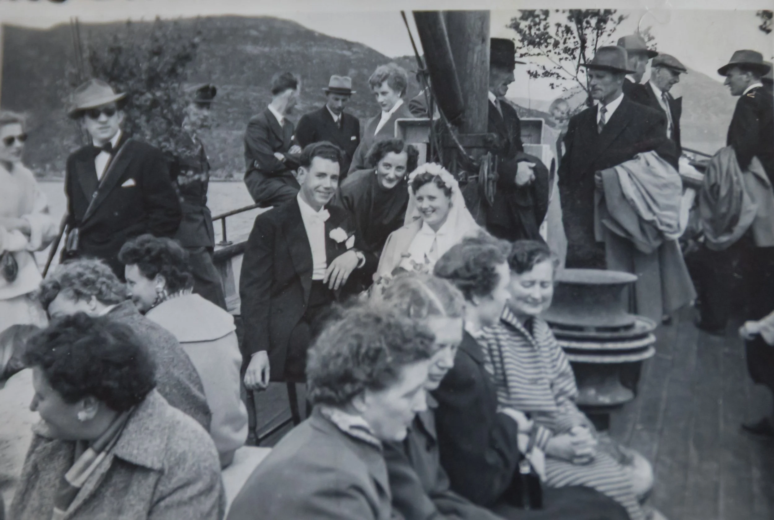 Oddrun Husevåg og Leif Sunde sammen med familie og bryllupsgjester om bord på fiskekutteren «Hovdenes» den 16. juni 1956. Brudeparet sitter omgitt av festkledde gjester på dekket. Fiskekutteren fraktet bryllupsfølget til vielsen i Måløy.