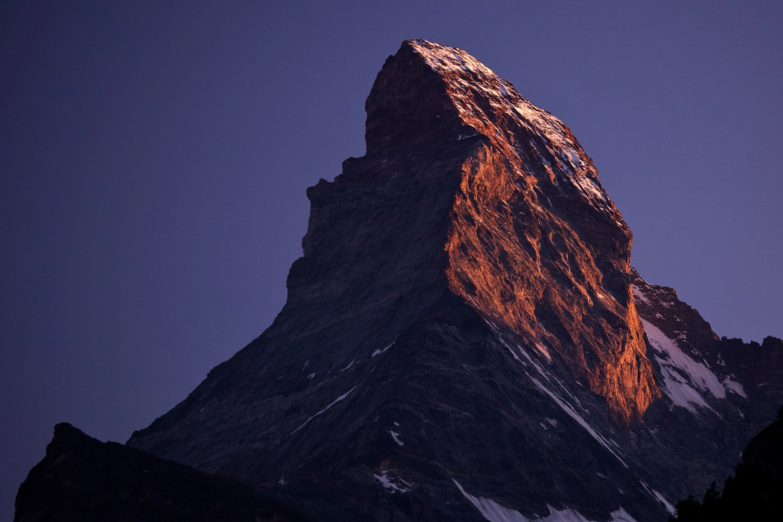 Warm evening light glowing on the steep face of the Matterhorn against a deep blue sky above the village of Zermatt.