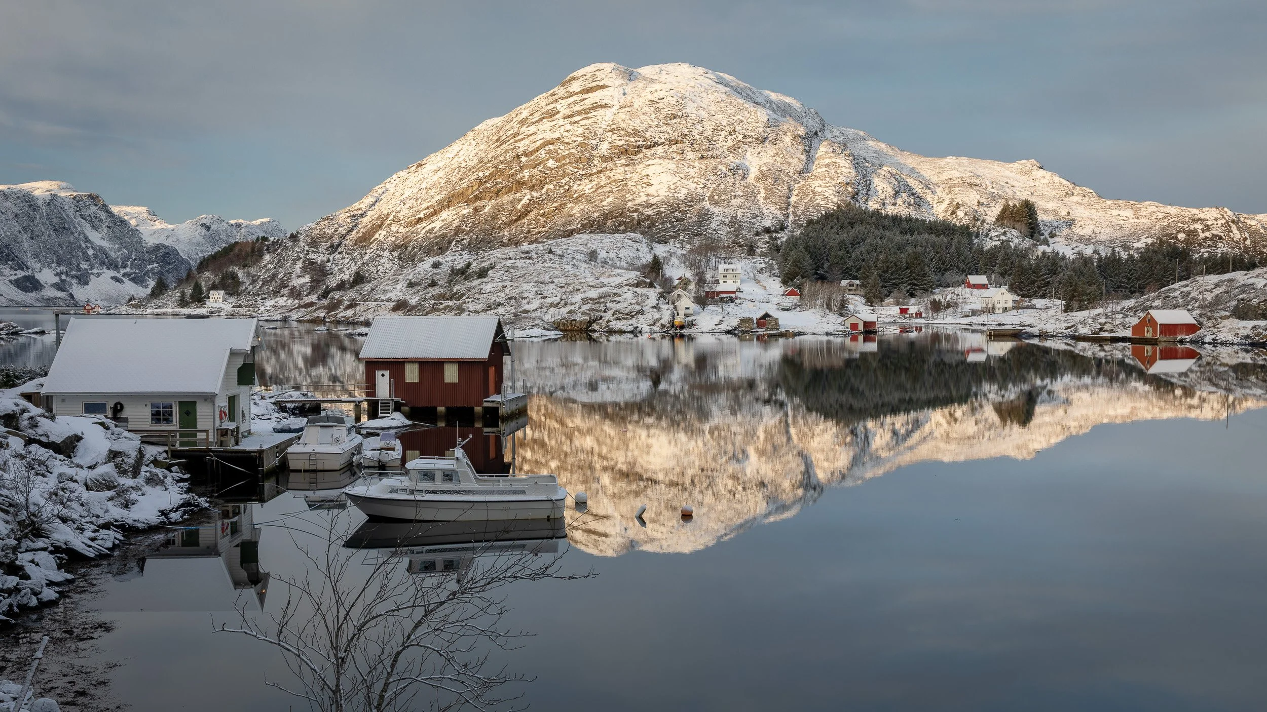 Winter light over Vågen on Husevågøy, Norway, with snow-covered shoreline, small boats, still water and soft reflections creating a quiet coastal landscape.