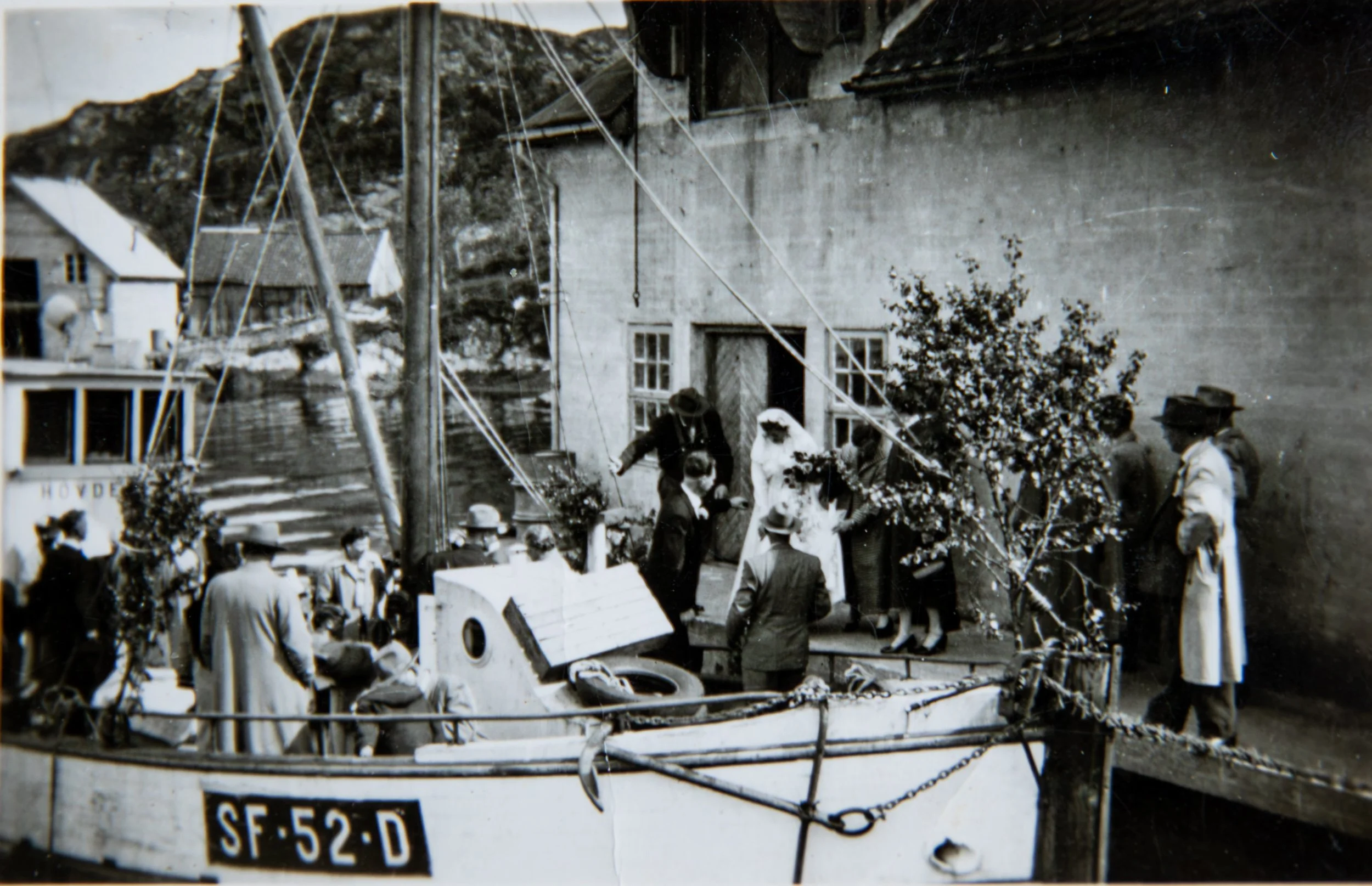 Historisk svart-hvitt fotografi fra 16. juni 1956. Bryllupsgjester samlet ved fiskekutteren «Hovdenes» (SF-52-D) i forbindelse med bryllupet til Oddrun Husevåg og Leif Sunde. Bruden i hvitt og flere festkledde gjester står ved kaien og fiskebåten.
