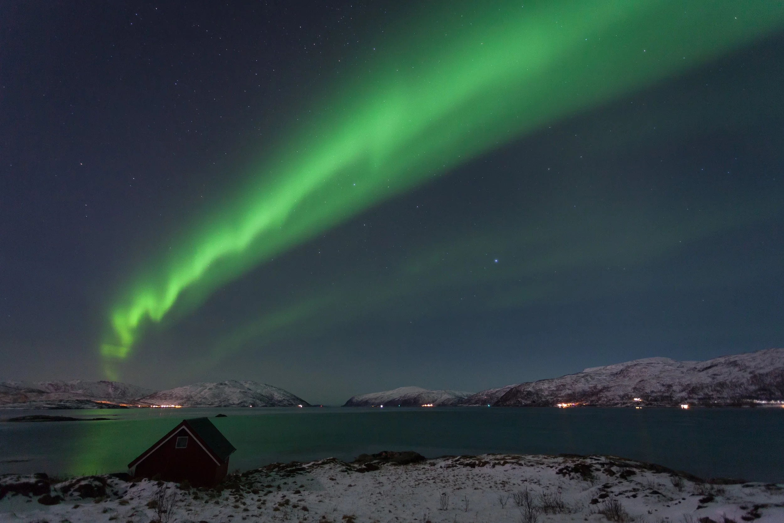 Northern Lights over Kvaløysundet near Tromsø, Norway – a bright green aurora ribbon stretches across the Arctic sky above a quiet fjord and a lone red cabin on the snowy shoreline.