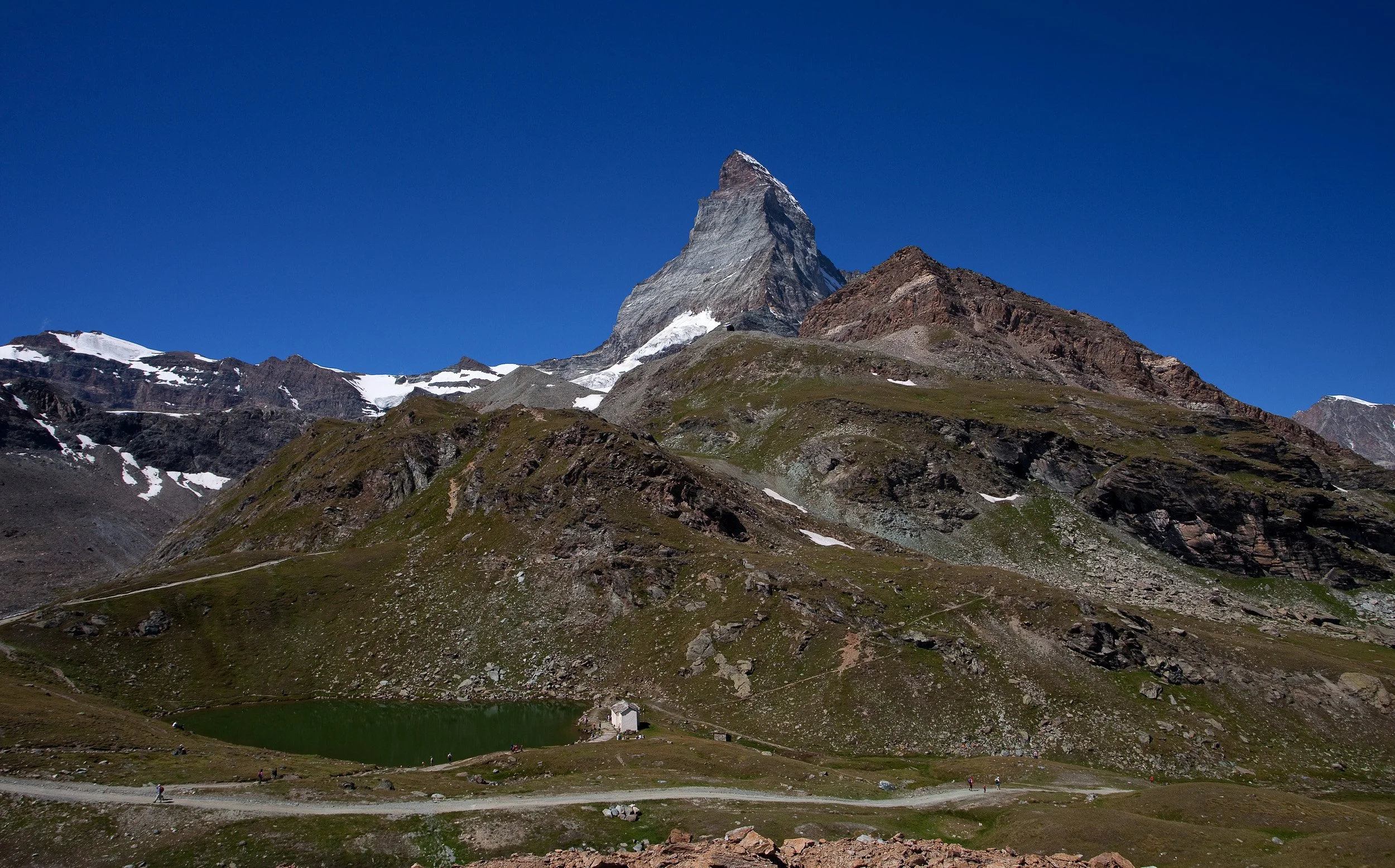 Matterhorn rising above rolling green ridges and a small mountain lake under a flawless high-summer sky in the Swiss Alps.