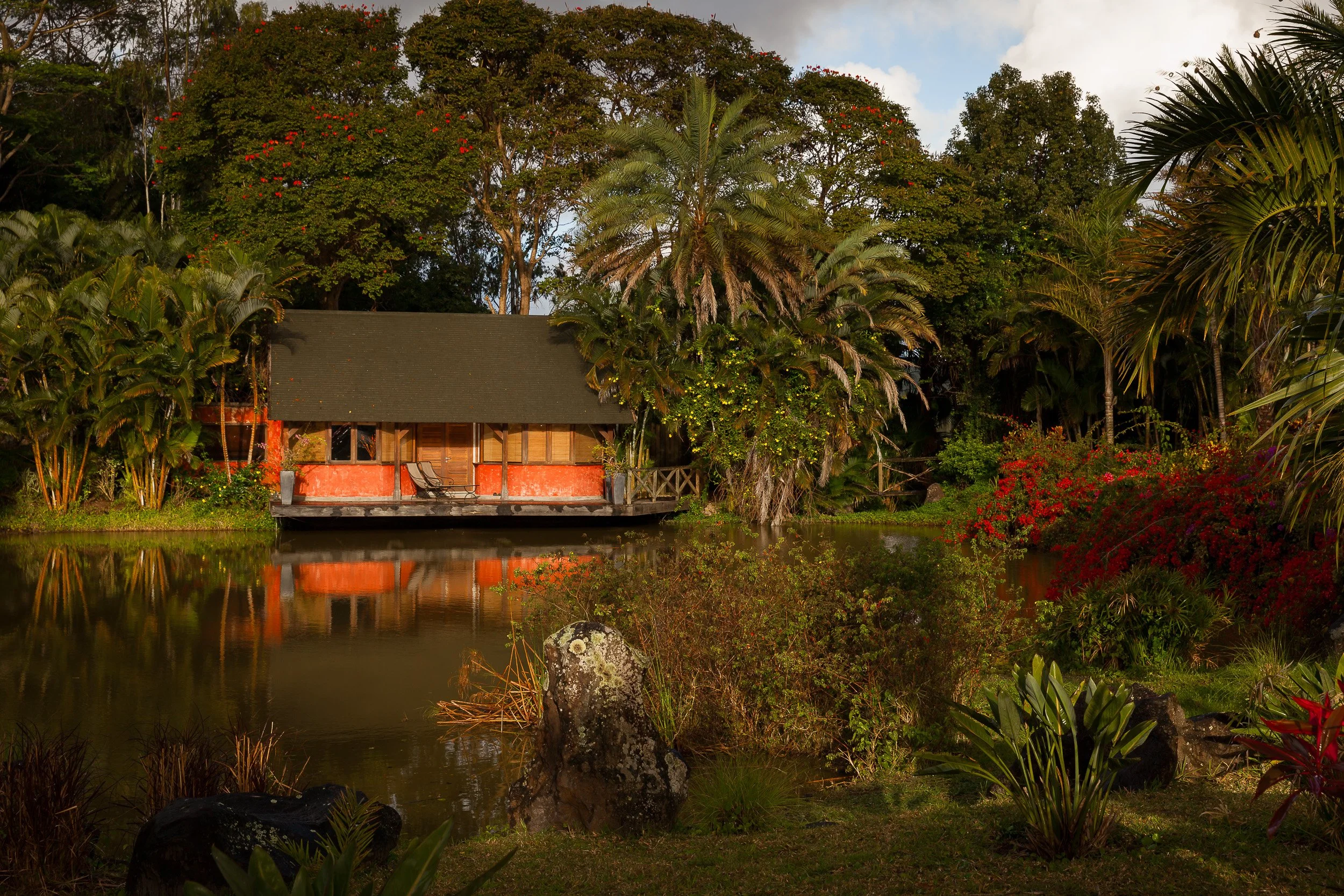 Secluded lakeside cabin at Lakaz Chamarel, its red walls and veranda reflected in still water and framed by lush palms and flowering bushes.