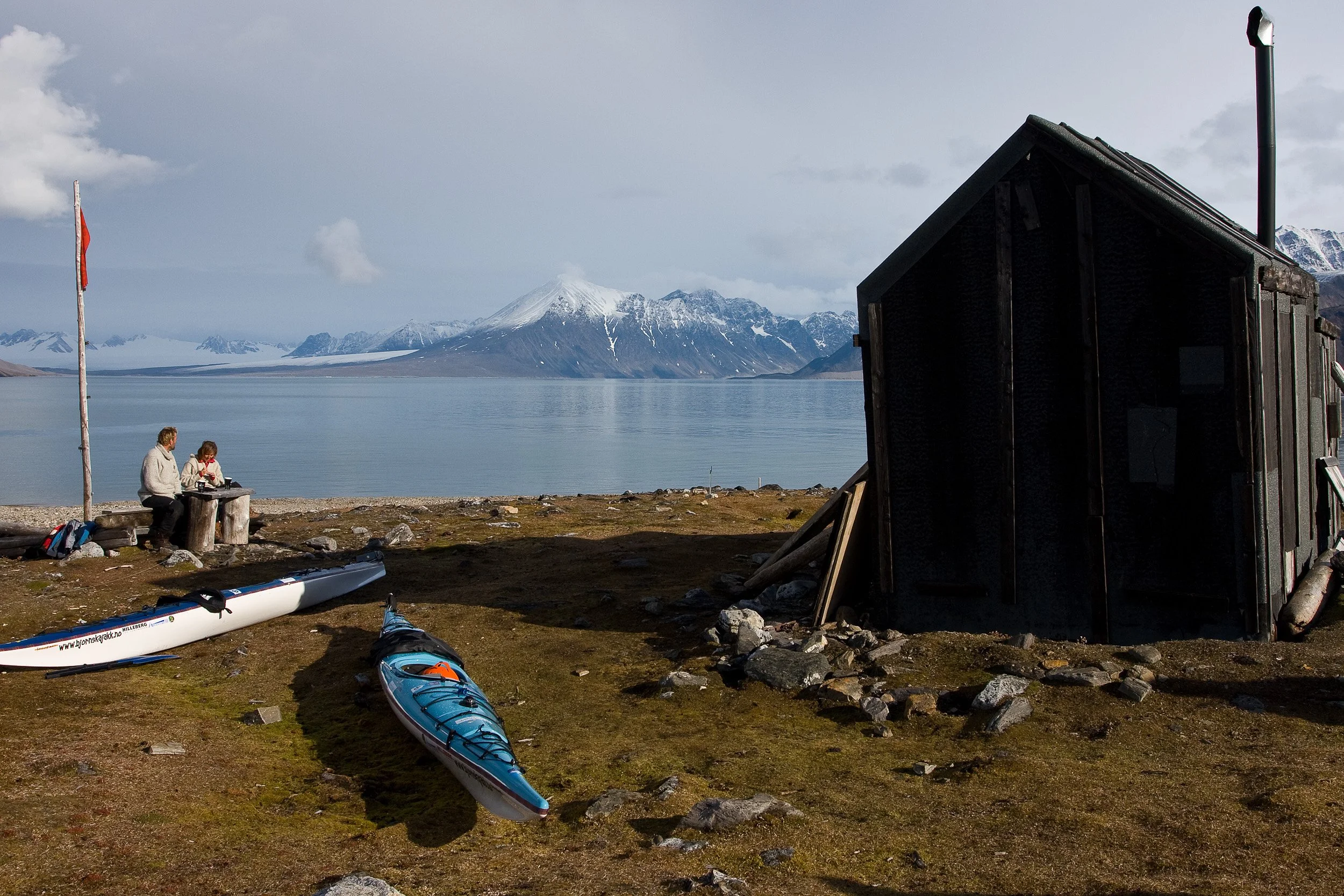 A simple hut and sea kayaks stand by the shore in Krossfjorden, facing calm water and snow-dusted mountains.