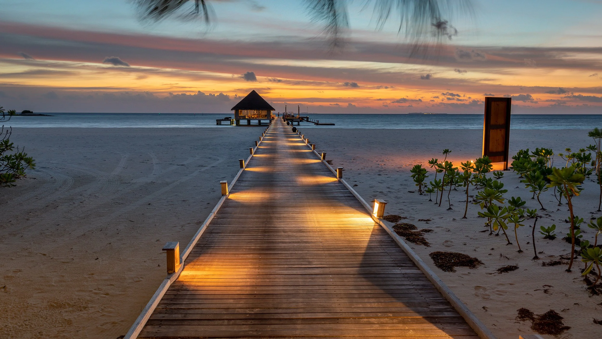 Sunset at Kanuhura in the Maldives, with a wooden jetty stretching across the lagoon towards warm evening light and soft tropical colour on the horizon.