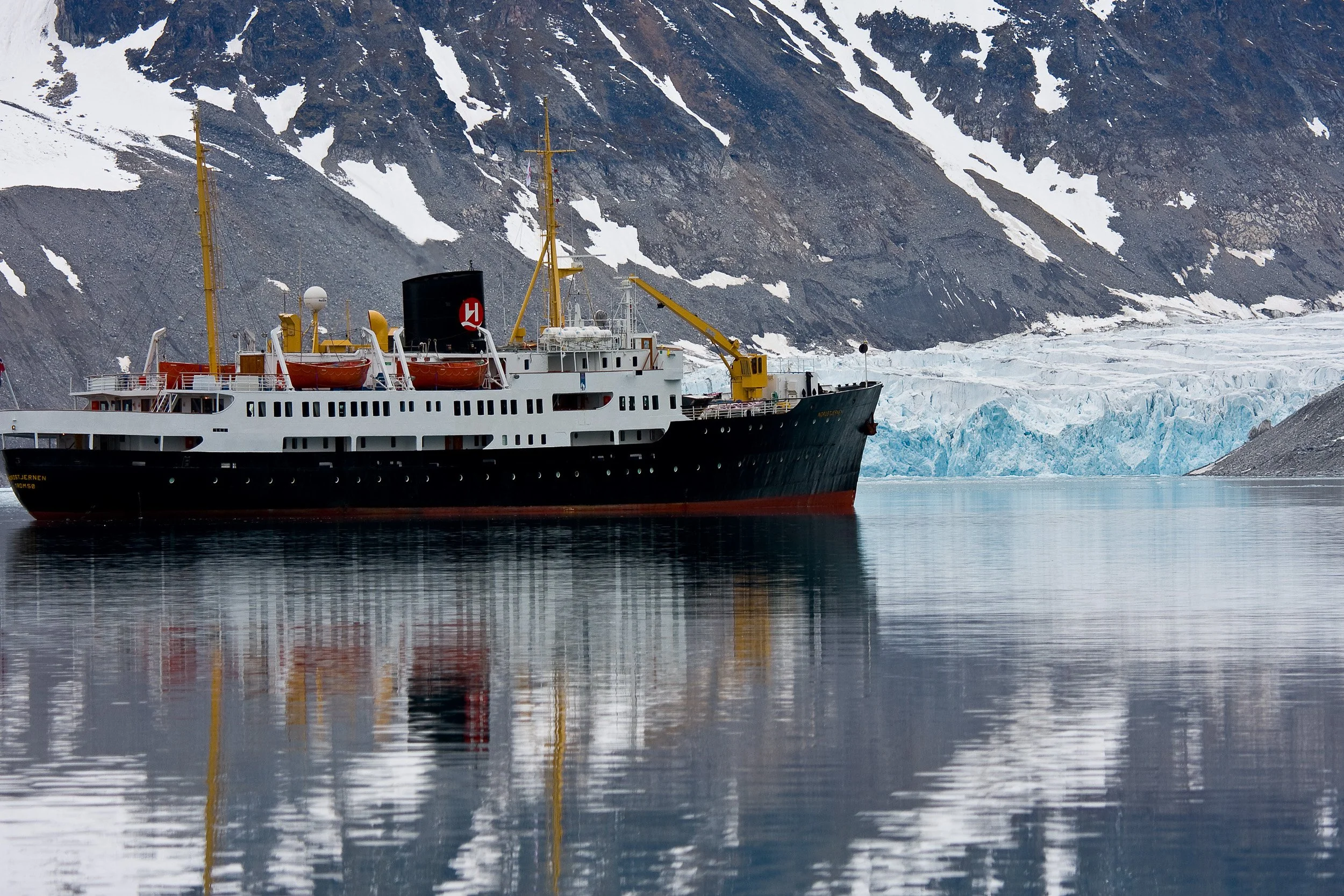 The expedition ship lies still beside glacier ice in Magdalenefjorden, mirrored in calm water.