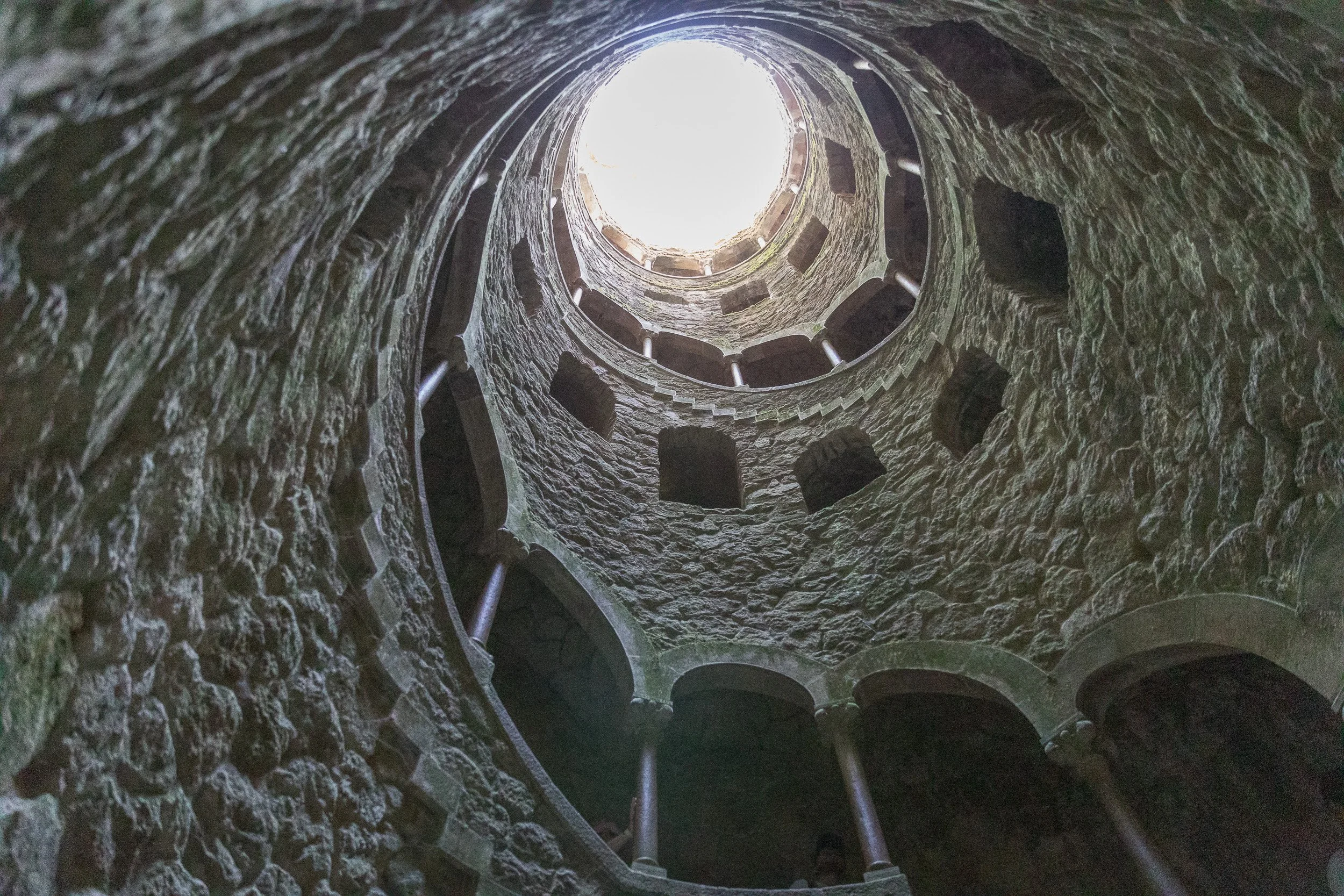 The Initiation Well at Quinta da Regaleira, seen from below as rough stone walls spiral upwards towards the daylight.