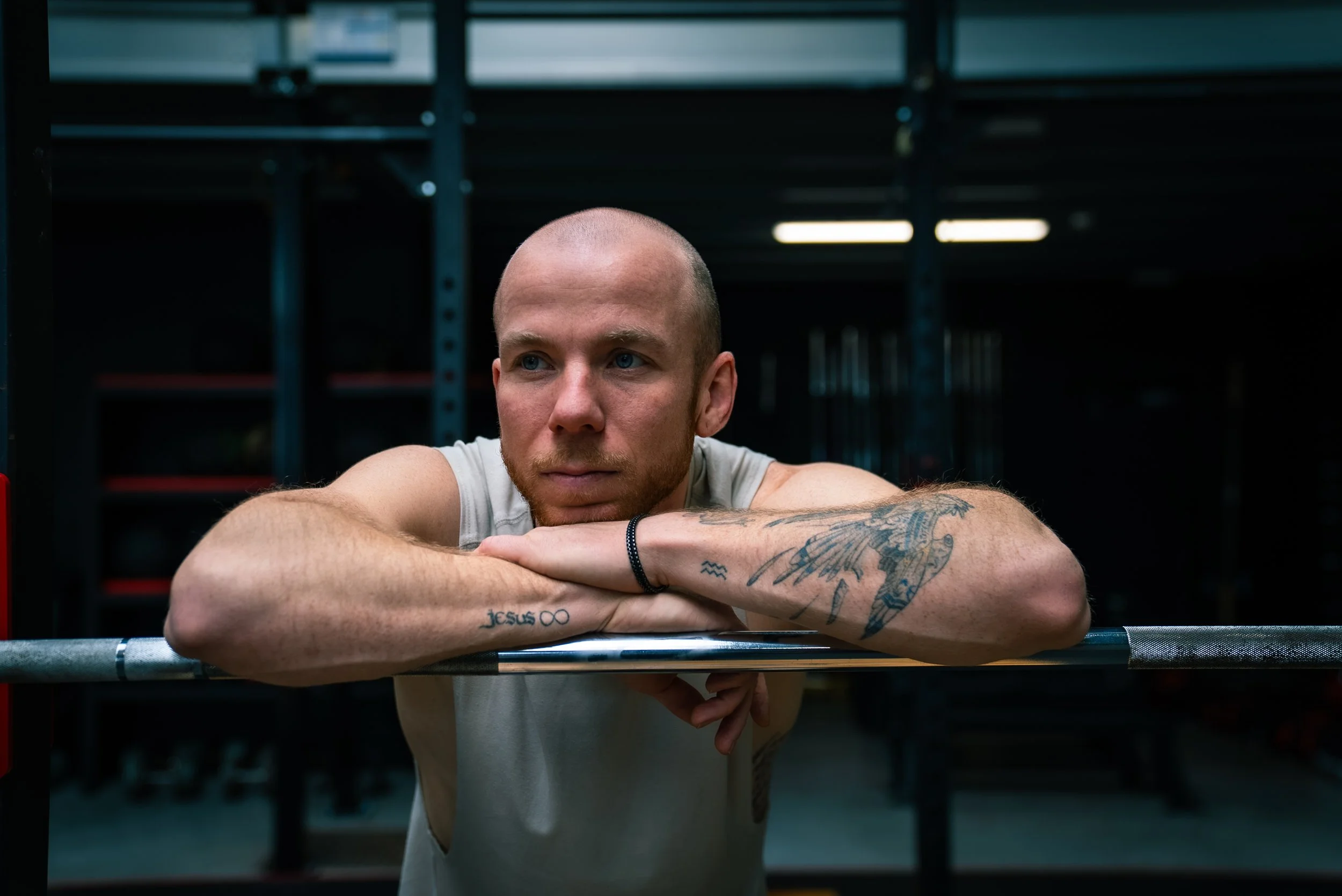A young man with tattoos, a shaved head, and a serious expression, resting his arms on a barbell in a gym.