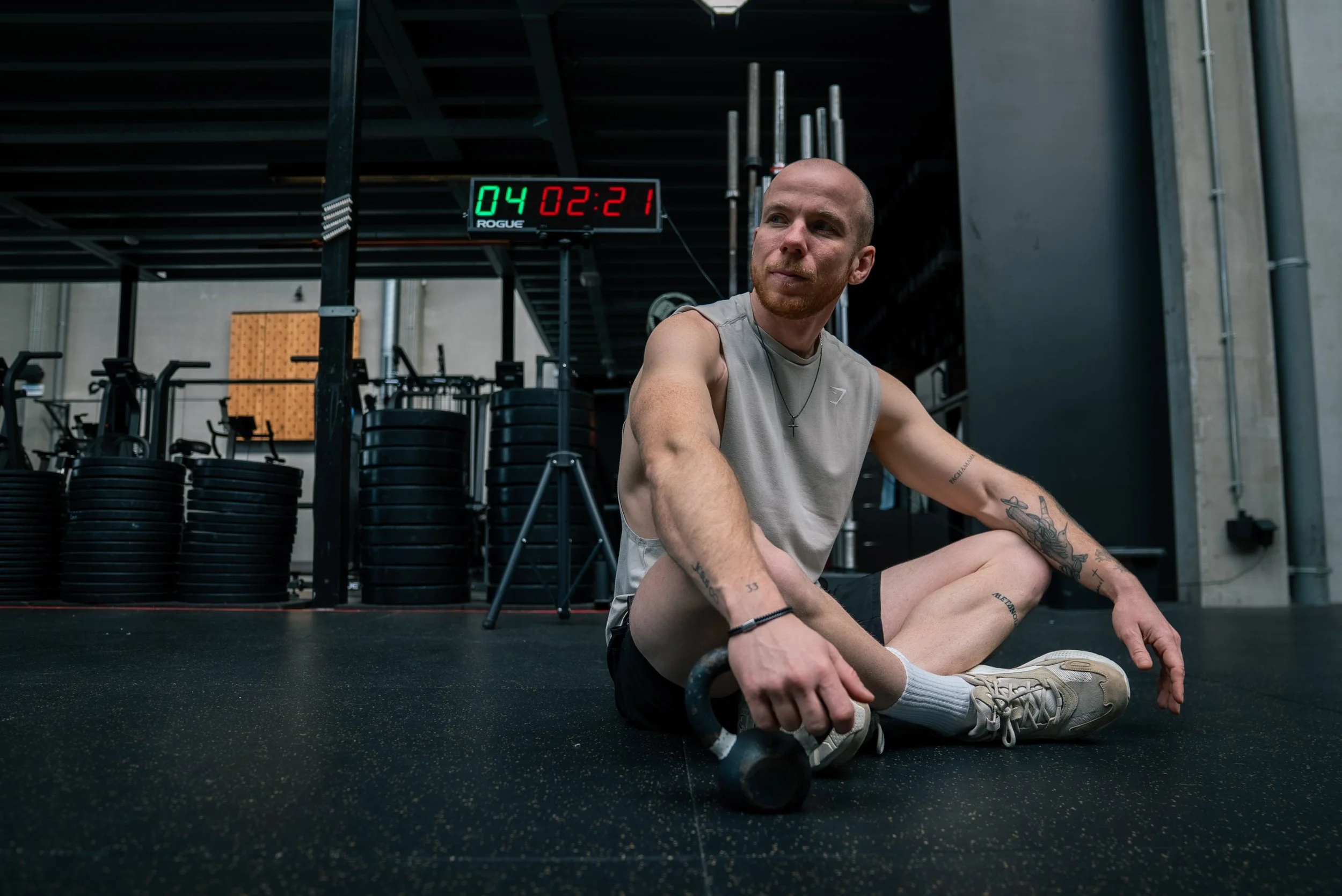 A man sitting on gym floor with kettlebell in hand after workout, in a gym with weight plates and digital scoreboard in background.