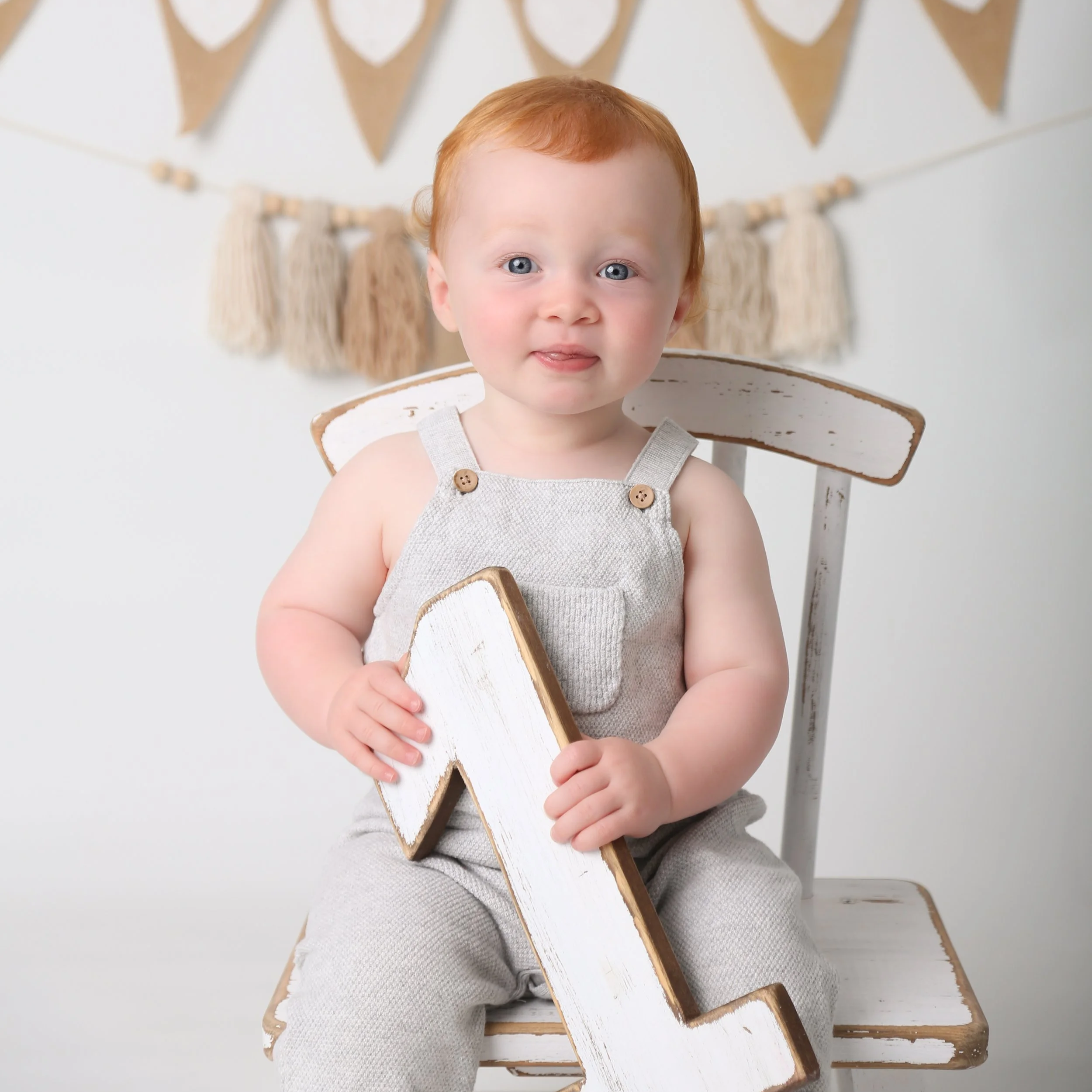 One-year-old on a white chair holding a wooden number one, cake smash photography in Upminster