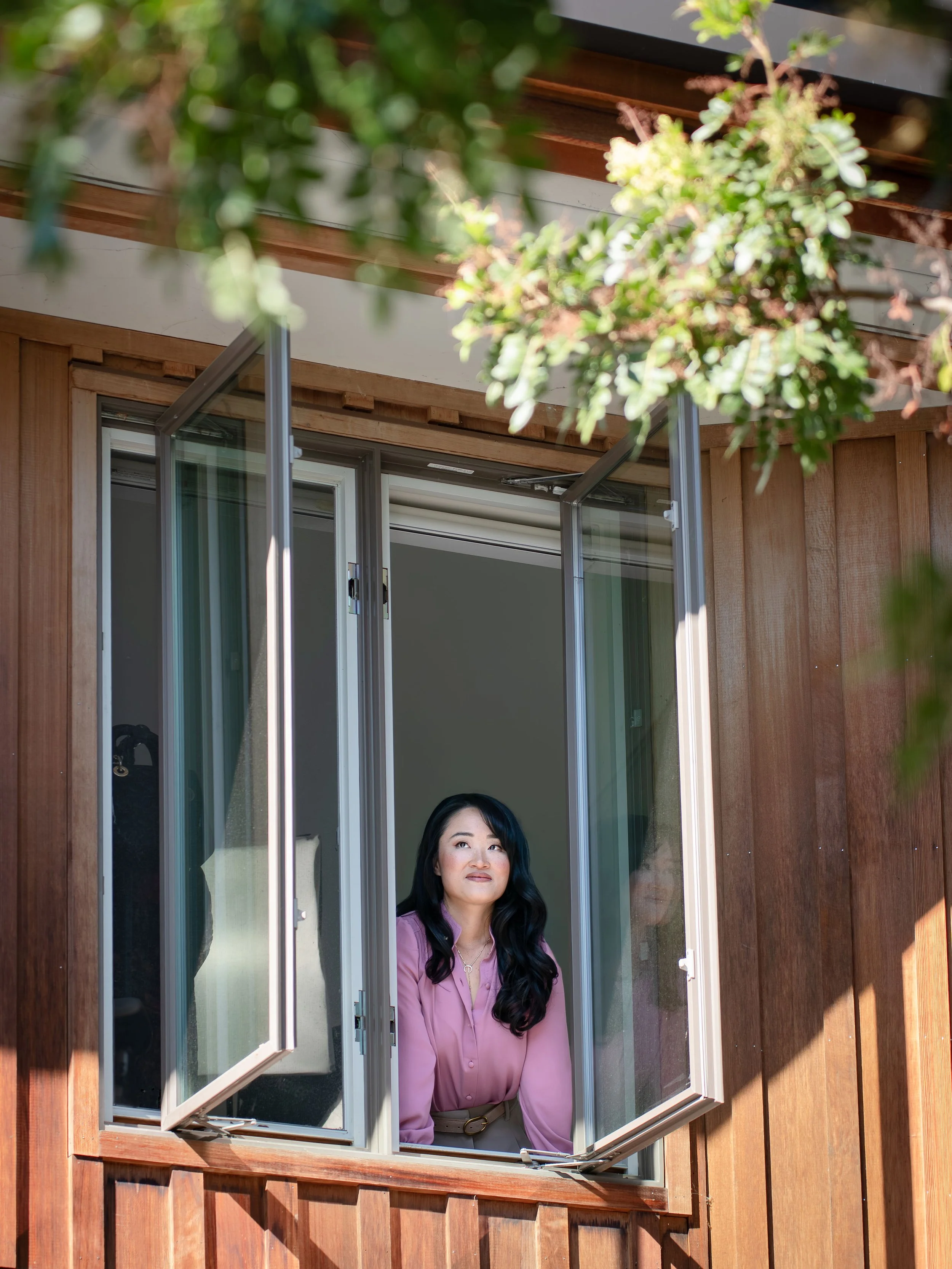 Elizabeth Huang with long black hair wearing a pink shirt looking out of a window of a wooden house.