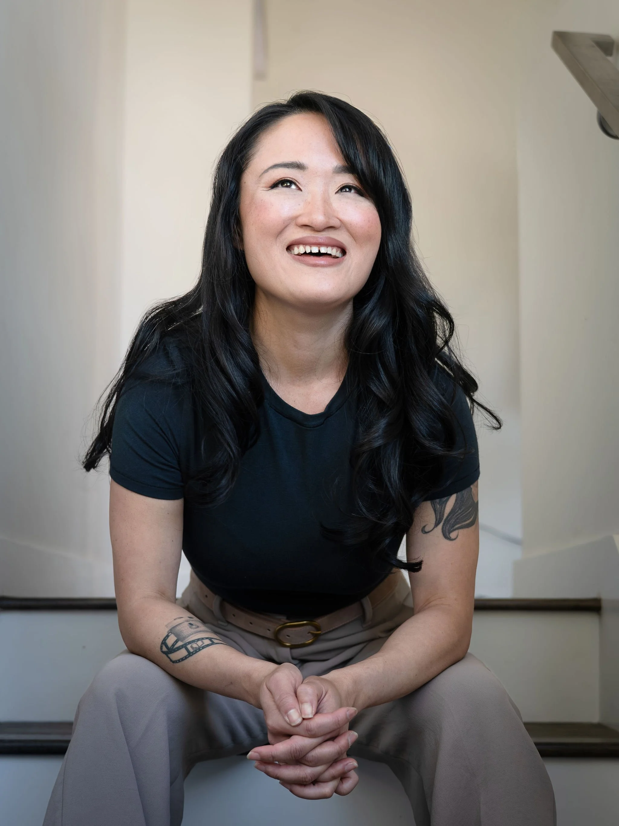 Elizabeth Huang with long black hair and tattoos on her arms, smiling and sitting on stairs.