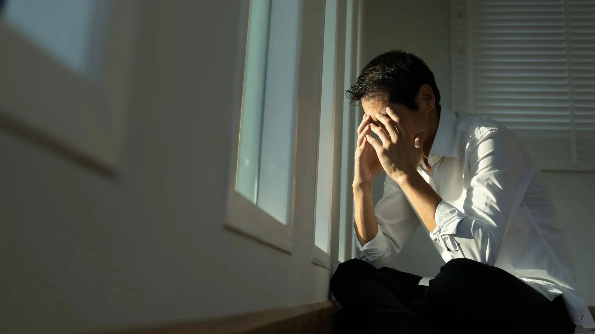 A man sitting on the floor by a window, holding his head in his hands, appearing distressed or overwhelmed.