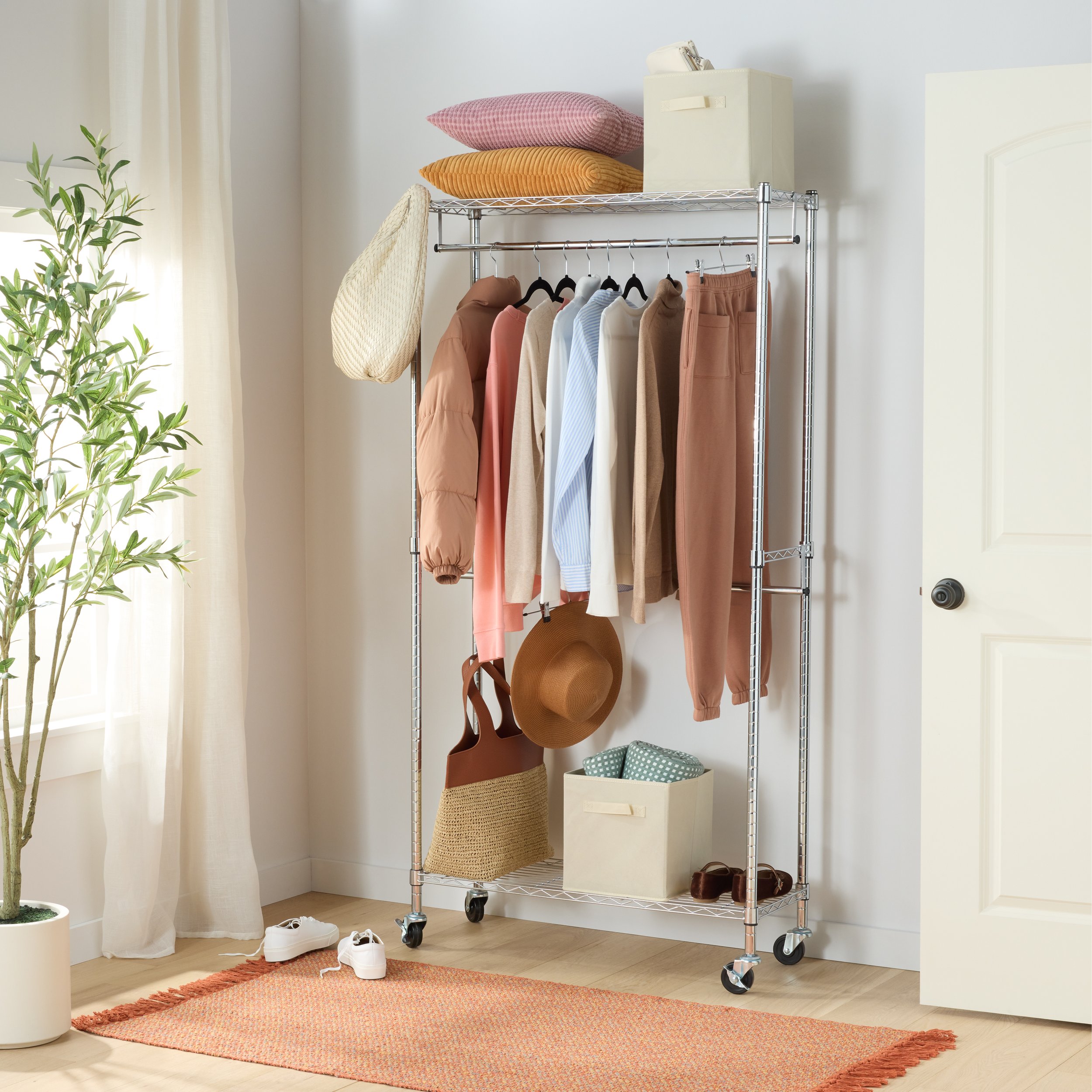 A metal clothing rack on wheels holding various clothing items such as shirts, jackets, and pants, with hats and storage boxes placed on top and underneath, located in a well-lit room with a potted plant, sneakers on the floor, and a door on the righ