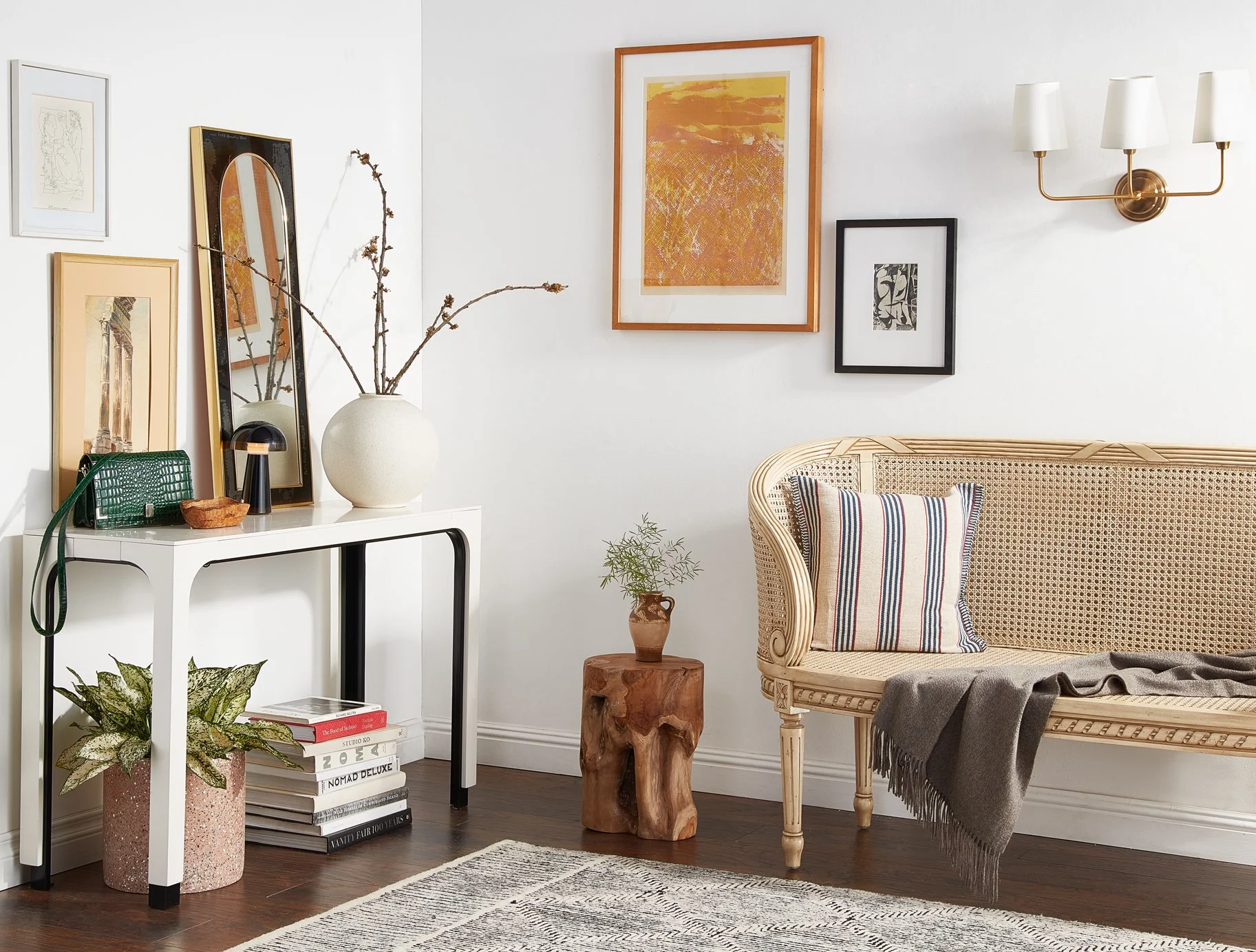 Living room corner with a white wall decorated with framed artwork and a gold wall sconce. A vintage-style cane sofa with a striped pillow and gray throw blanket. A natural wood side table with green plant in a rustic pot. A white console table with 