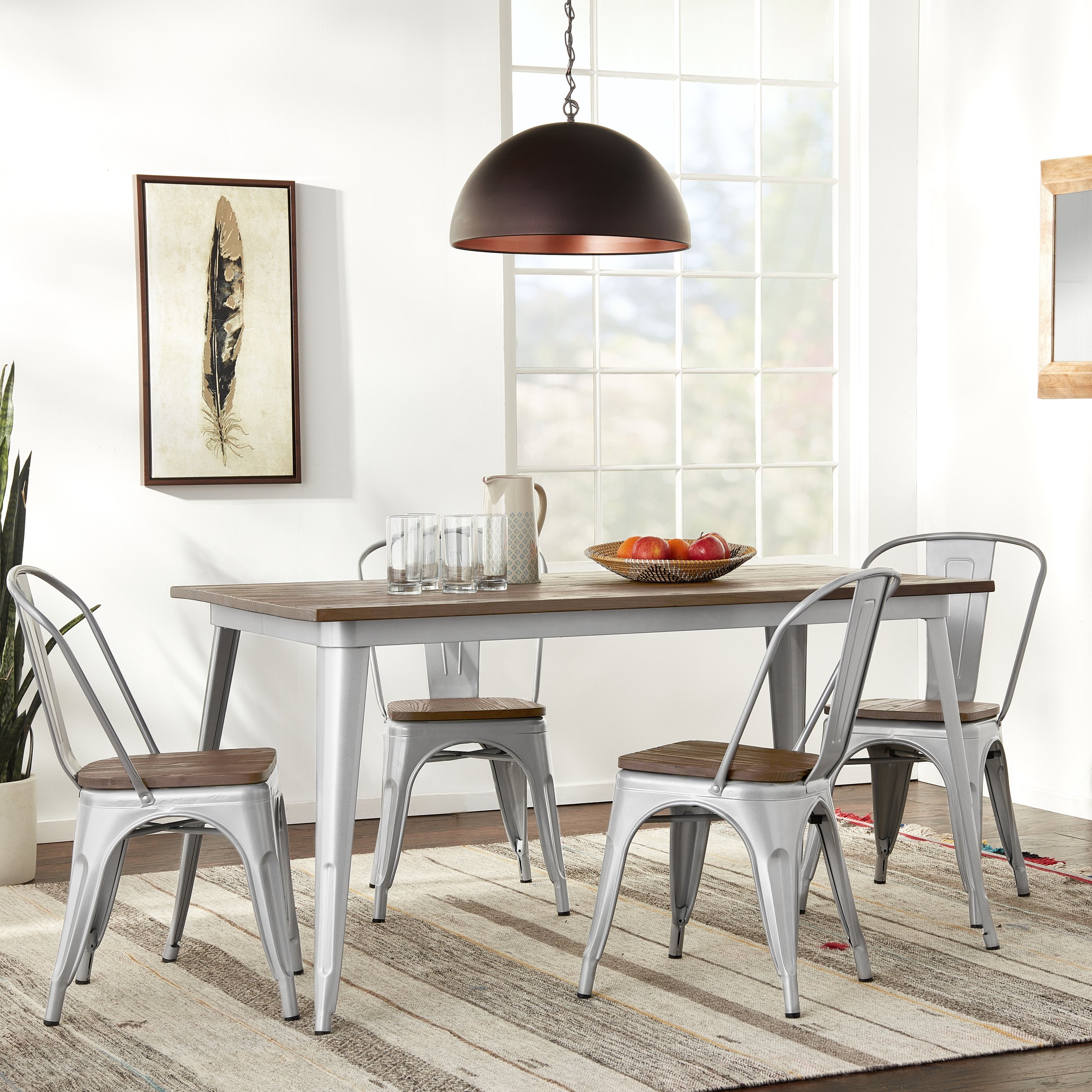 Modern dining room with a wooden table, metal chairs with wooden seats, a large black pendant light, a framed feather artwork on white wall, a pitcher, glasses, a bowl of fruit, and natural light coming through large windows.