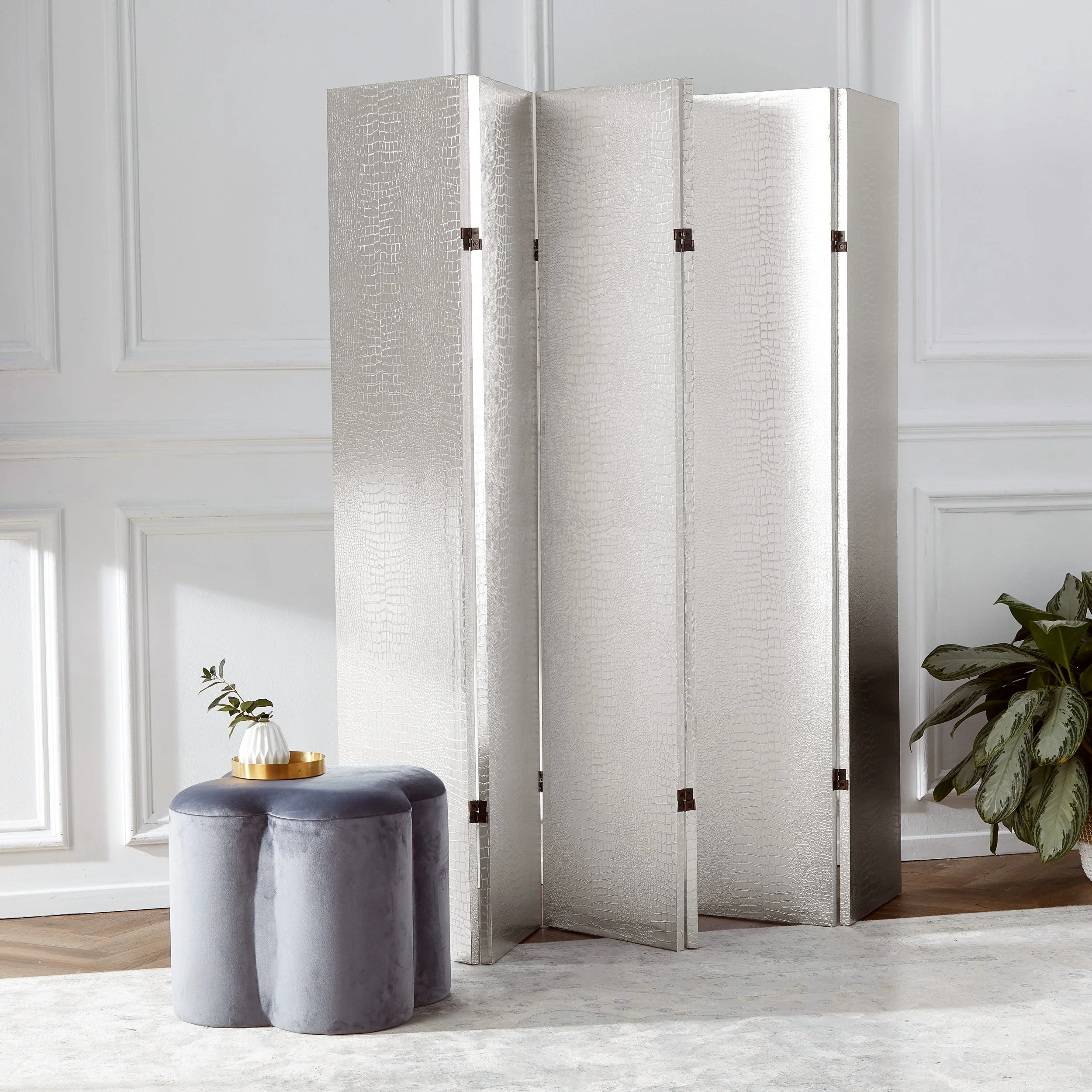 A silver room divider with a crocodile skin pattern, beside a small gray velvet stool with a white vase and greenery, in a room with white panel walls and a light-colored rug.