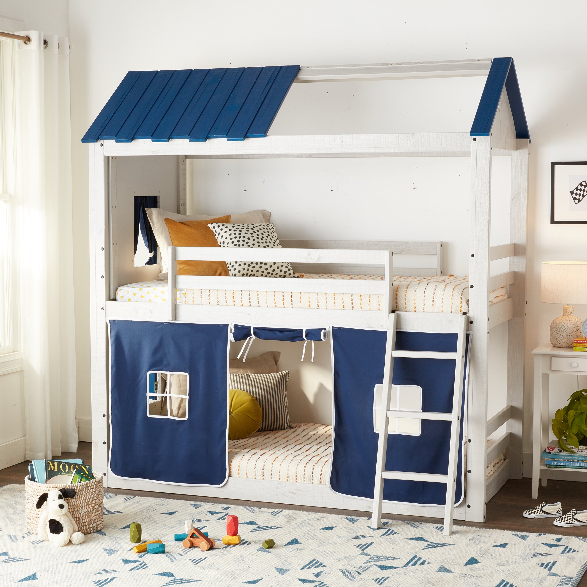 A bedroom with a white bunk bed that has a climbing ladder and a blue canopy on the top bunk. The bottom bunk has a blue curtain with a window opening. There are pillows on both bunks, and toys scattered on a blue and white rug. A basket with books a