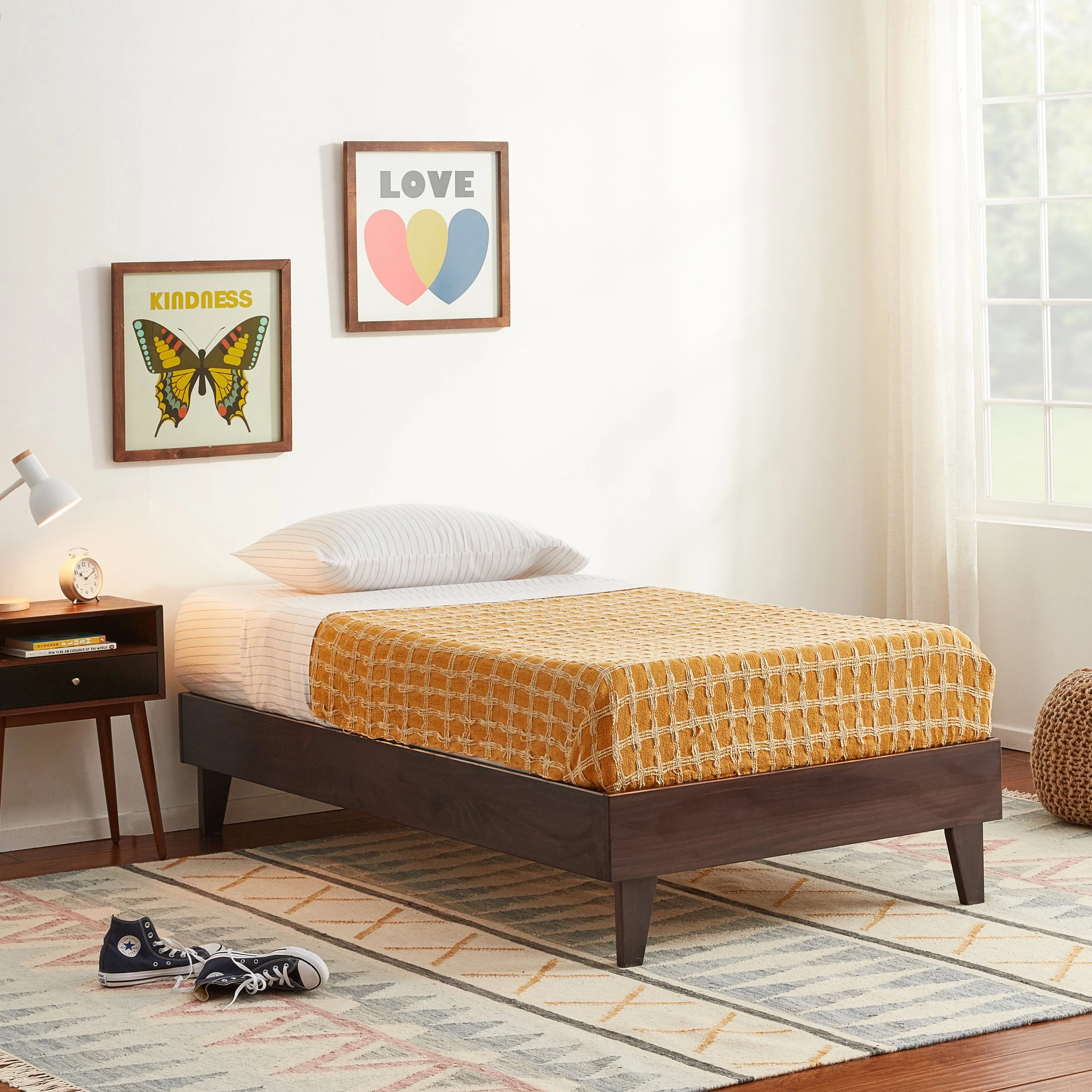 Cozy bedroom with a bed, orange checkered blanket, white pillow, large window with cream curtains, wall art with butterfly, love, kindness words, small bedside table, clock, books, and sneakers on a patterned rug.