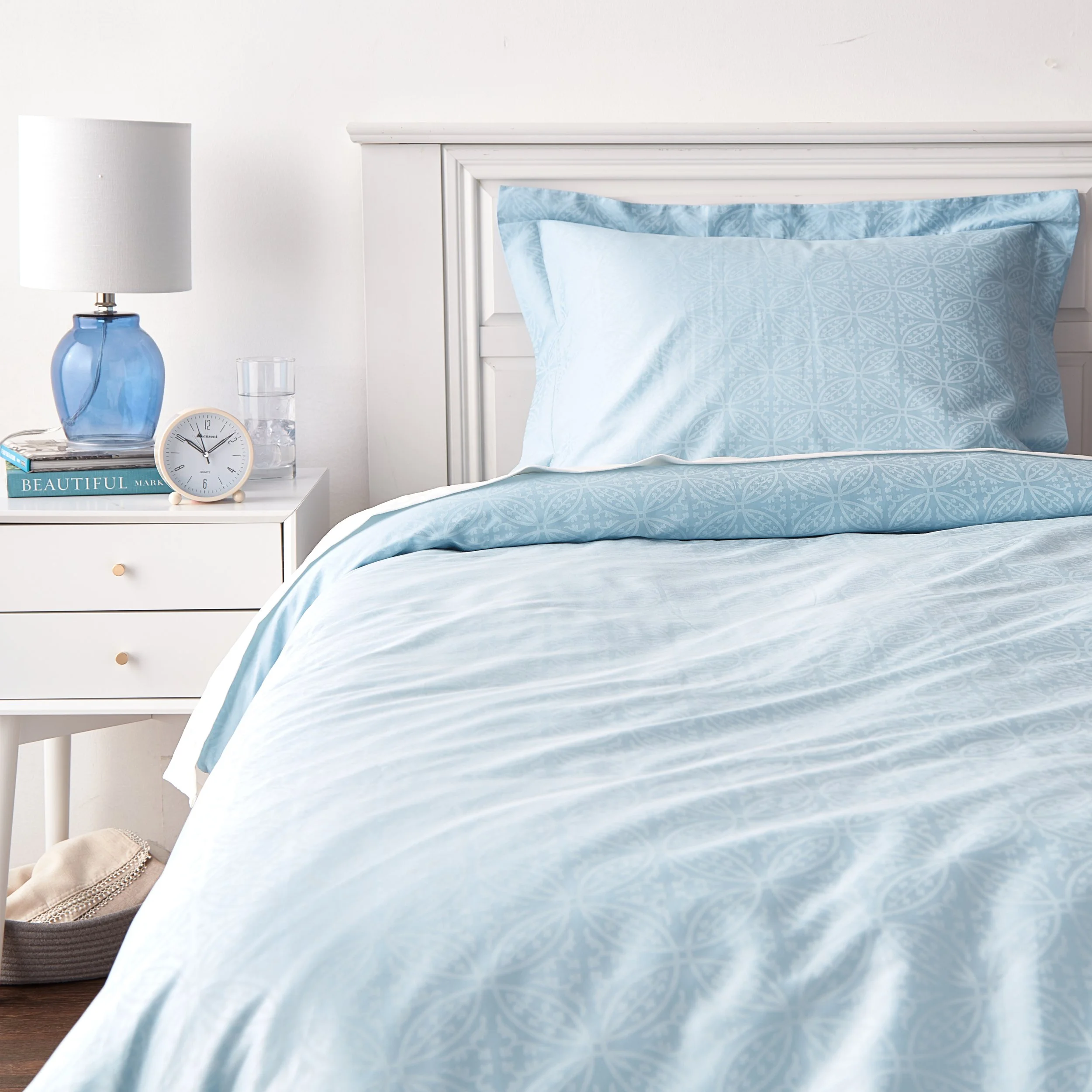 A neatly made bed with light blue patterned bedding and pillows, a white headboard, and a white nightstand with a blue lamp, a small clock, a glass of water, and a book.