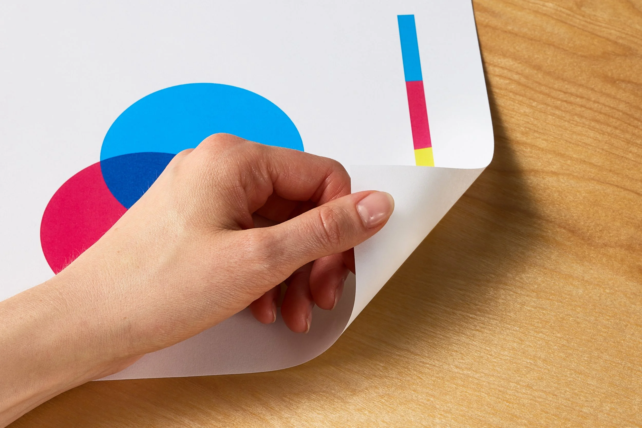 A person peeling back a corner of paper with overlapping blue and red circles and a color bar on white paper placed on a wooden surface.
