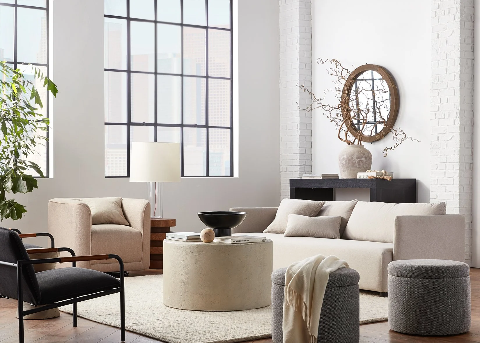 Living room with large windows, white walls, a cream-colored sofa, a black armchair with wooden arms, a white round coffee table with books and decor, a beige armchair with a side table, a black console table with decorative items and a round mirror,