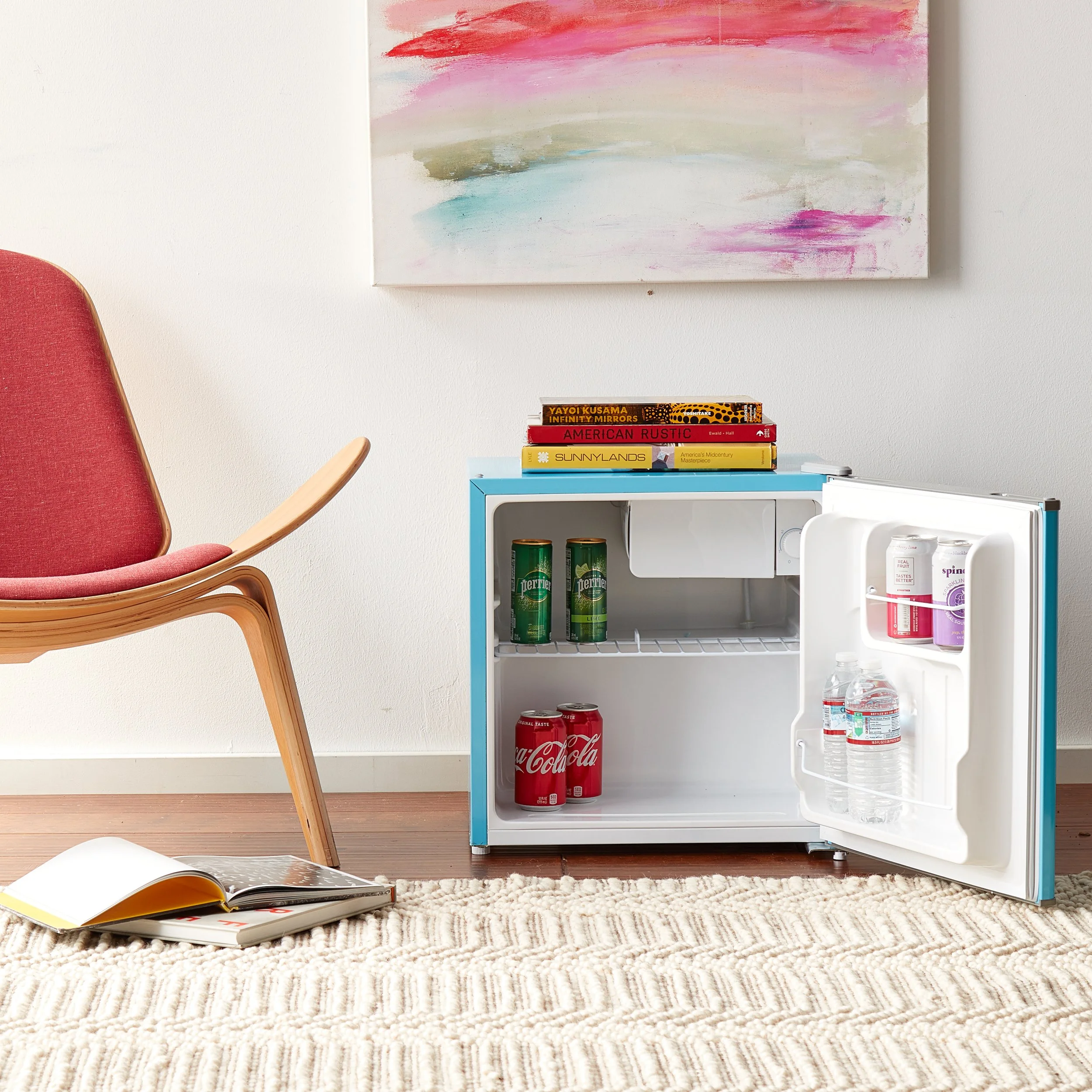 Small blue refrigerator with a few cans of drinks and bottled water, a stack of books on top, an open magazine on a striped rug, a pink and beige armchair, and an abstract pink, green, and white painting on the wall.