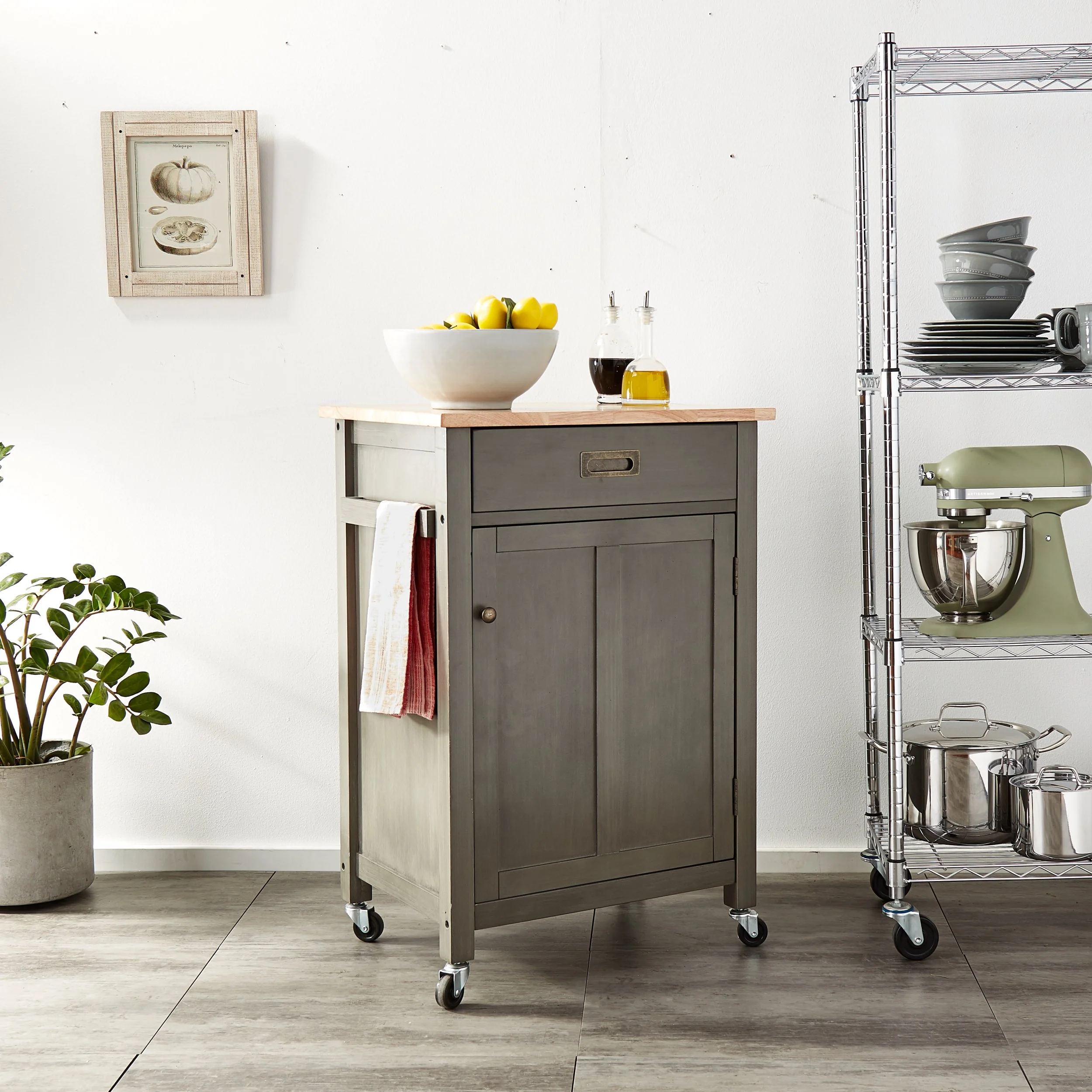 A kitchen corner with a gray rolling cabinet topped with a white bowl of lemons, oil and vinegar bottles, a potted plant, a wall picture of a pumpkin, and a metal shelving unit with bowls, mugs, and stainless steel cookware.