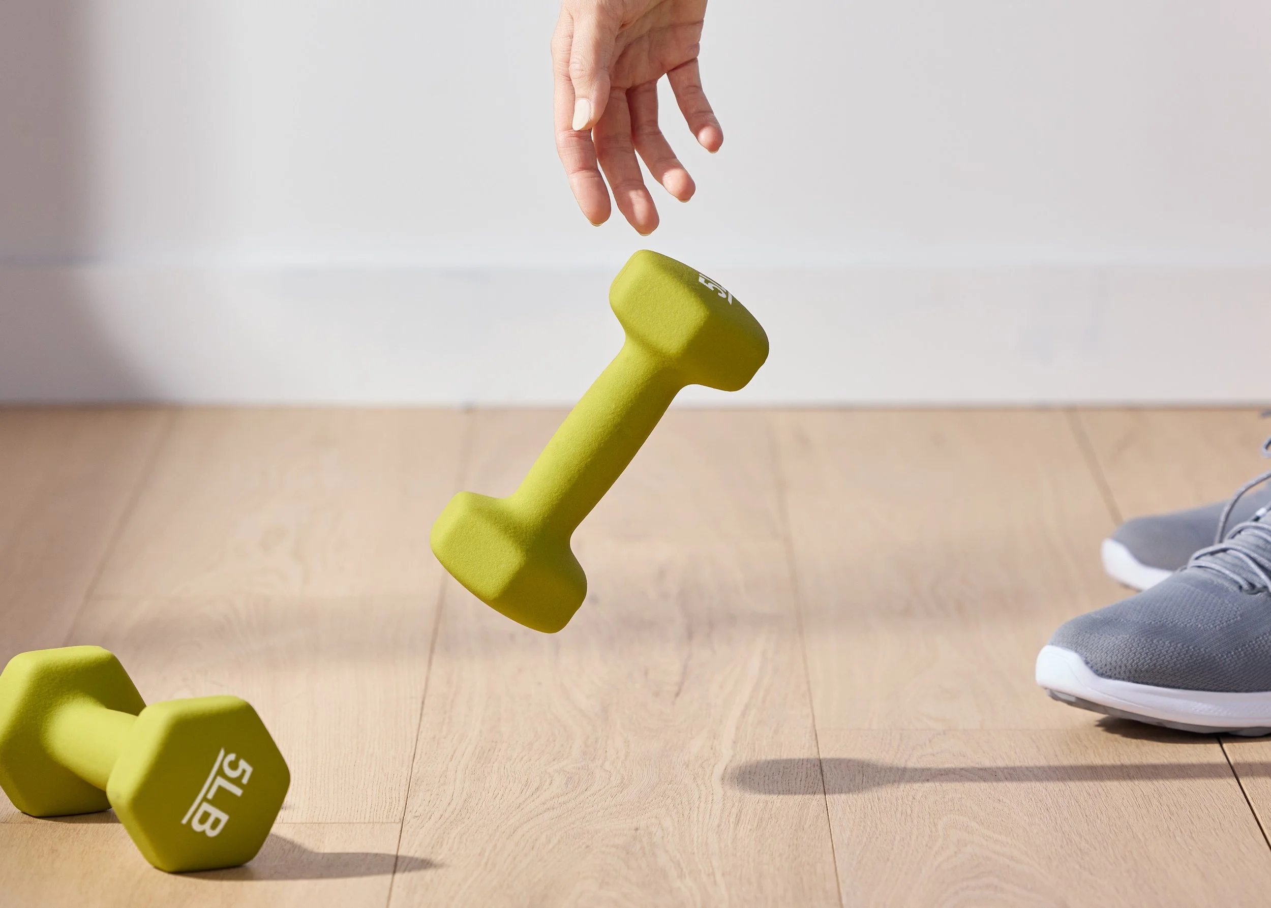 A person is dropping a yellow dumbbell while two other yellow dumbbells and a gray athletic shoe are on a wooden floor.