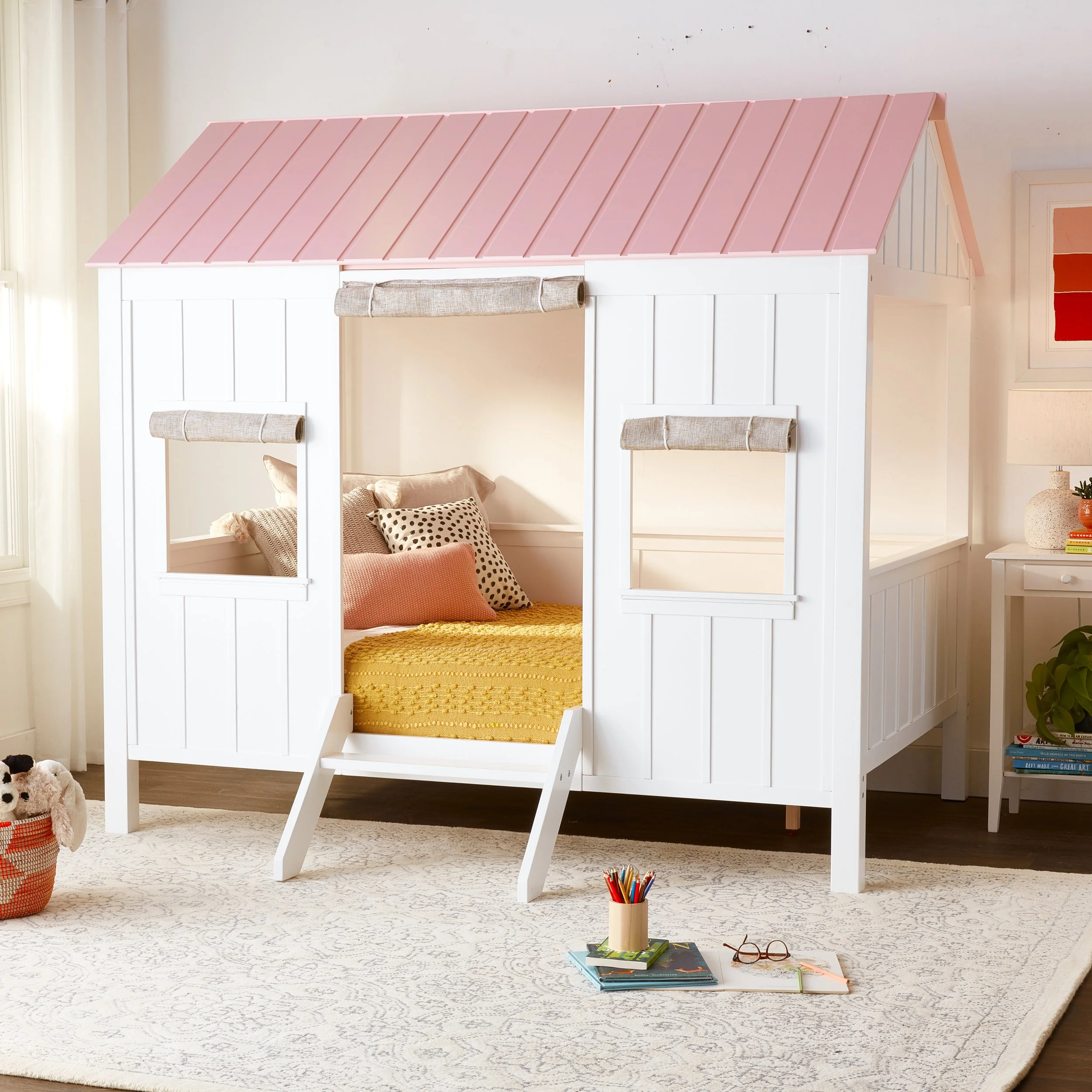 A white playhouse bed with a pink roof, inside a child's bedroom, with a cozy bed, pillows, and toys.