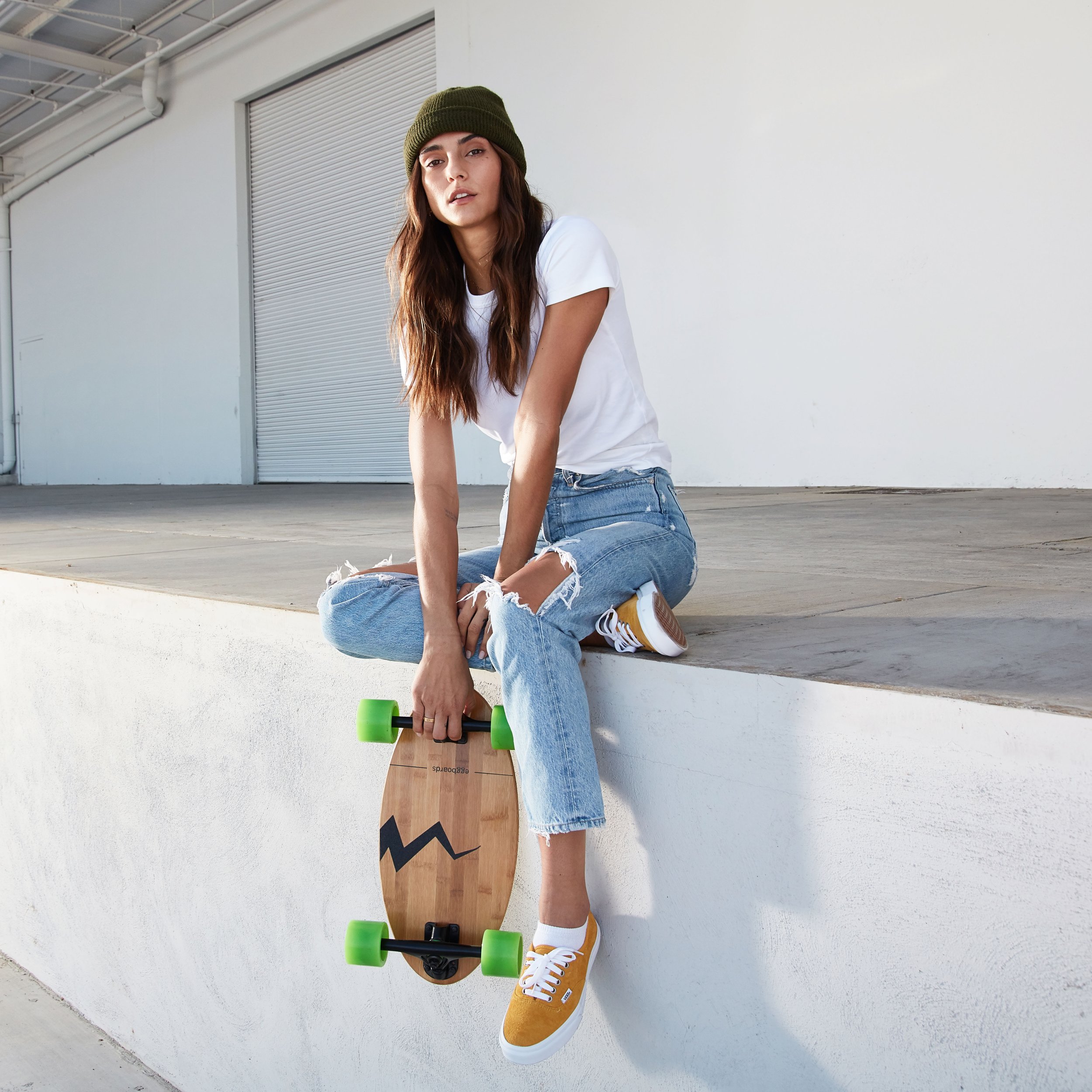 A young woman with long brown hair, wearing a green beanie, white t-shirt, ripped jeans, and yellow sneakers, sitting on a ledge with a skateboard hanging off her leg, holding a green skateboard with neon green wheels.