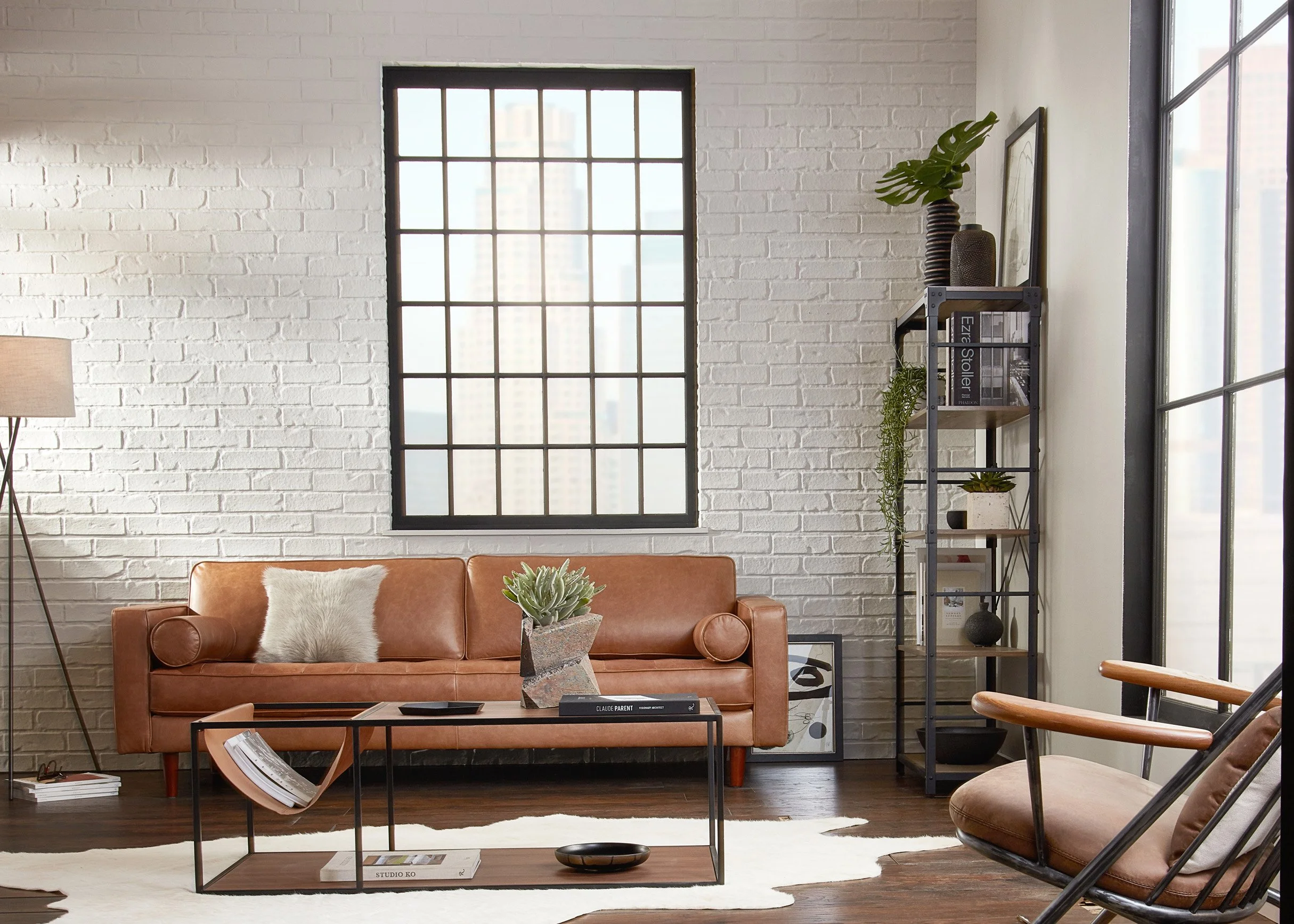 Modern living room with a leather sofa, a glass-top coffee table, a wooden armchair, a tall metal and wood bookshelf, large windows, and a white brick wall.