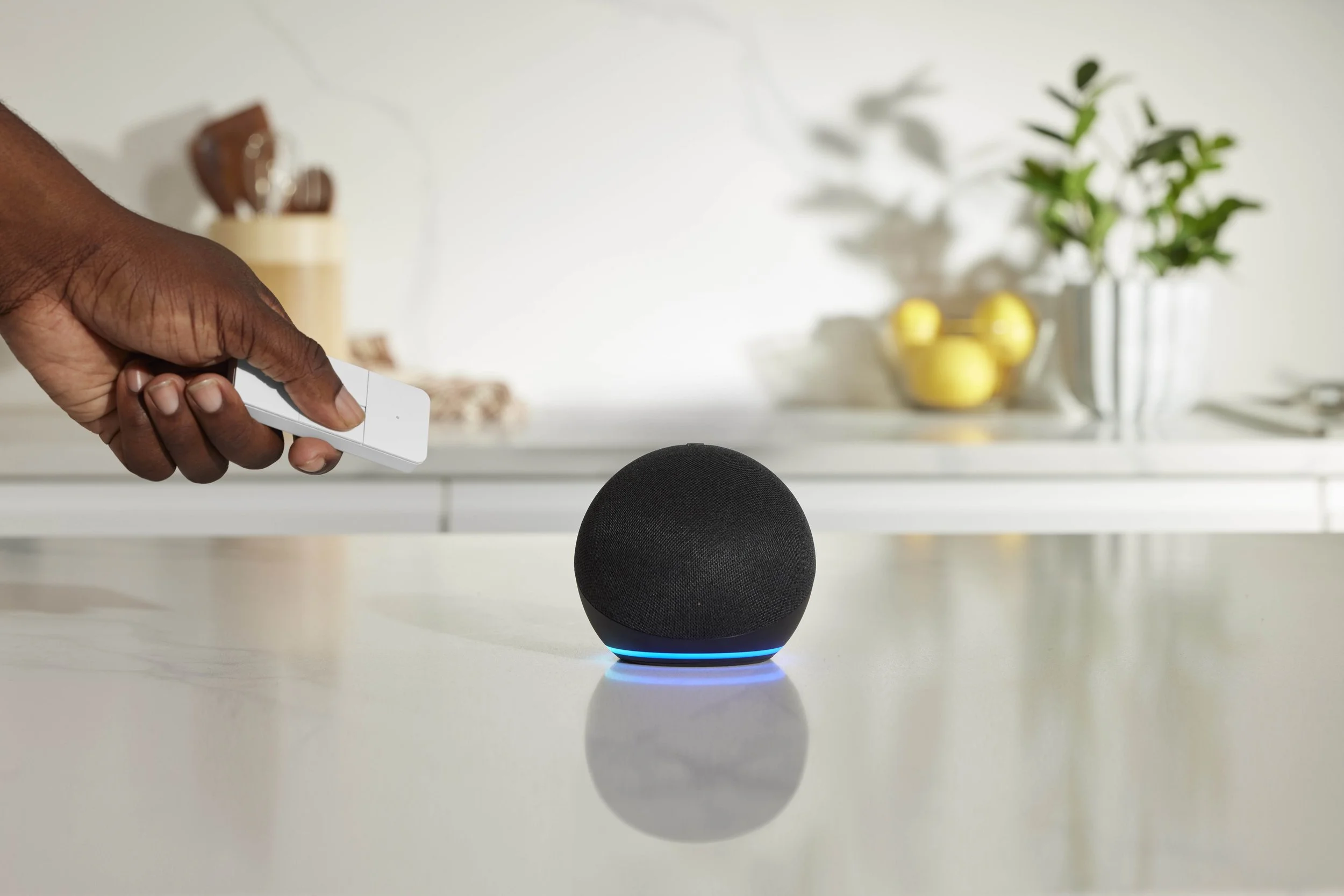 A person is holding a remote control pointing at a black smart speaker with a glowing blue ring placed on a white table.