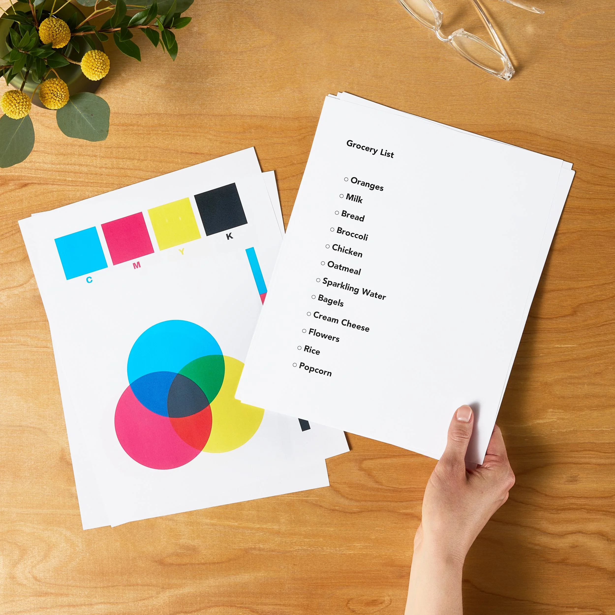 A person holding a grocery list printed on white paper, with other papers containing colorful diagrams and text on a wooden table. The grocery list includes items such as oranges, milk, bread, broccoli, chicken, oatmeal, sparkling water, bagels, cream cheese, flowers, rice, and popcorn. A small flower arrangement and clear glasses are also visible on the table.