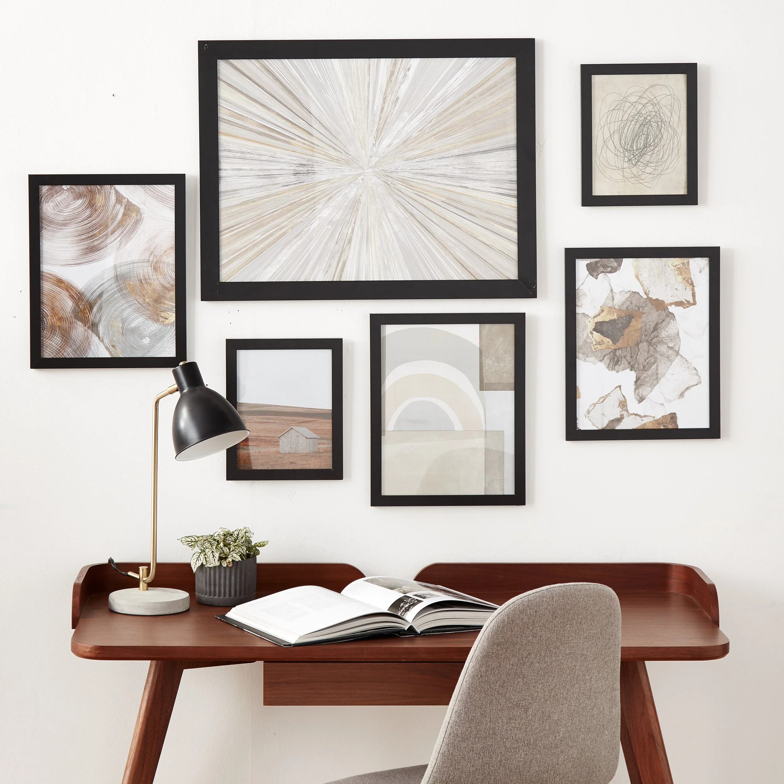 Home office with a wooden desk, open book, black desk lamp, gray chair, and wall art with abstract and nature-themed prints in black frames
