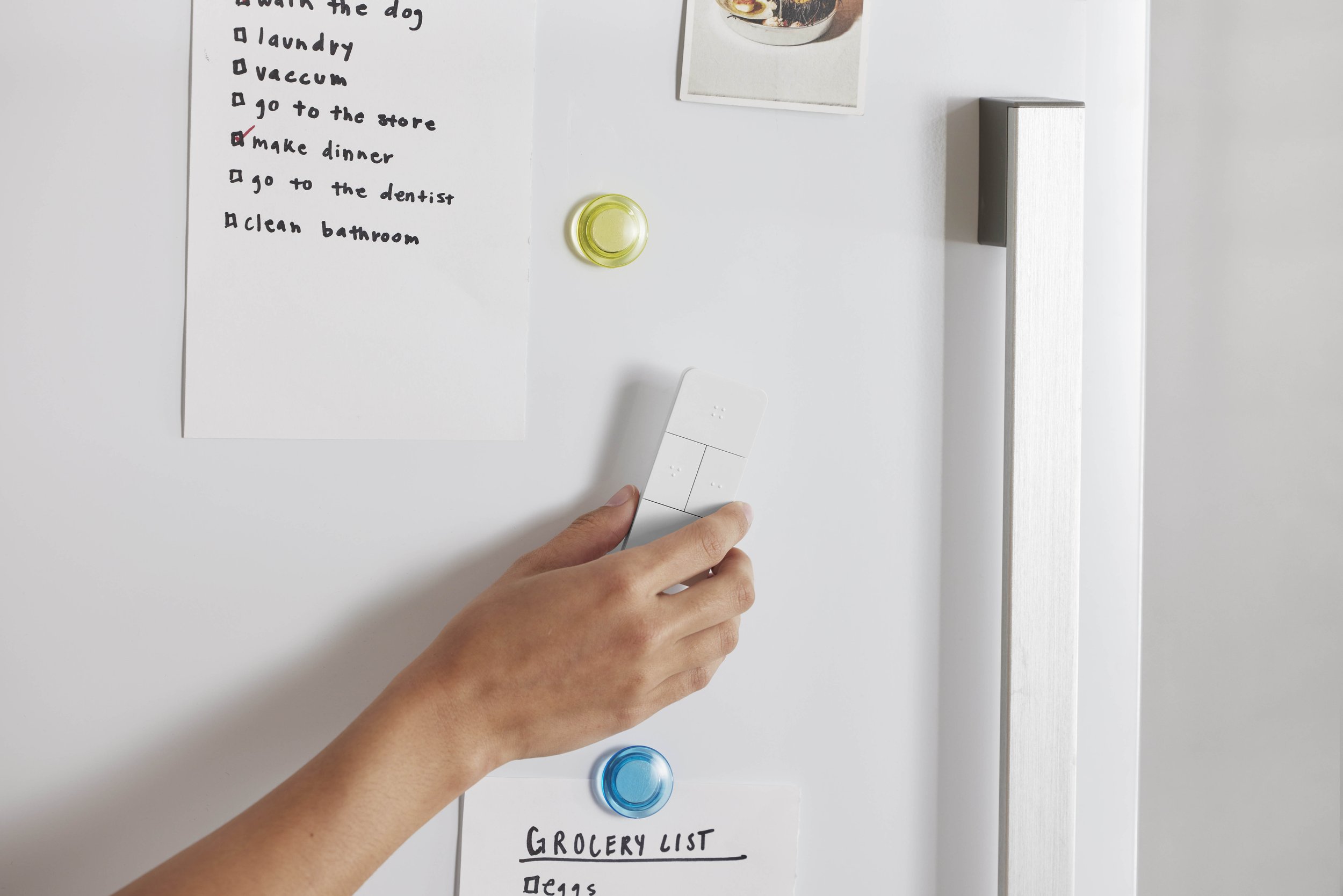 A hand holding a white remote control on a white refrigerator with grocery and to-do lists attached, and colorful magnets.