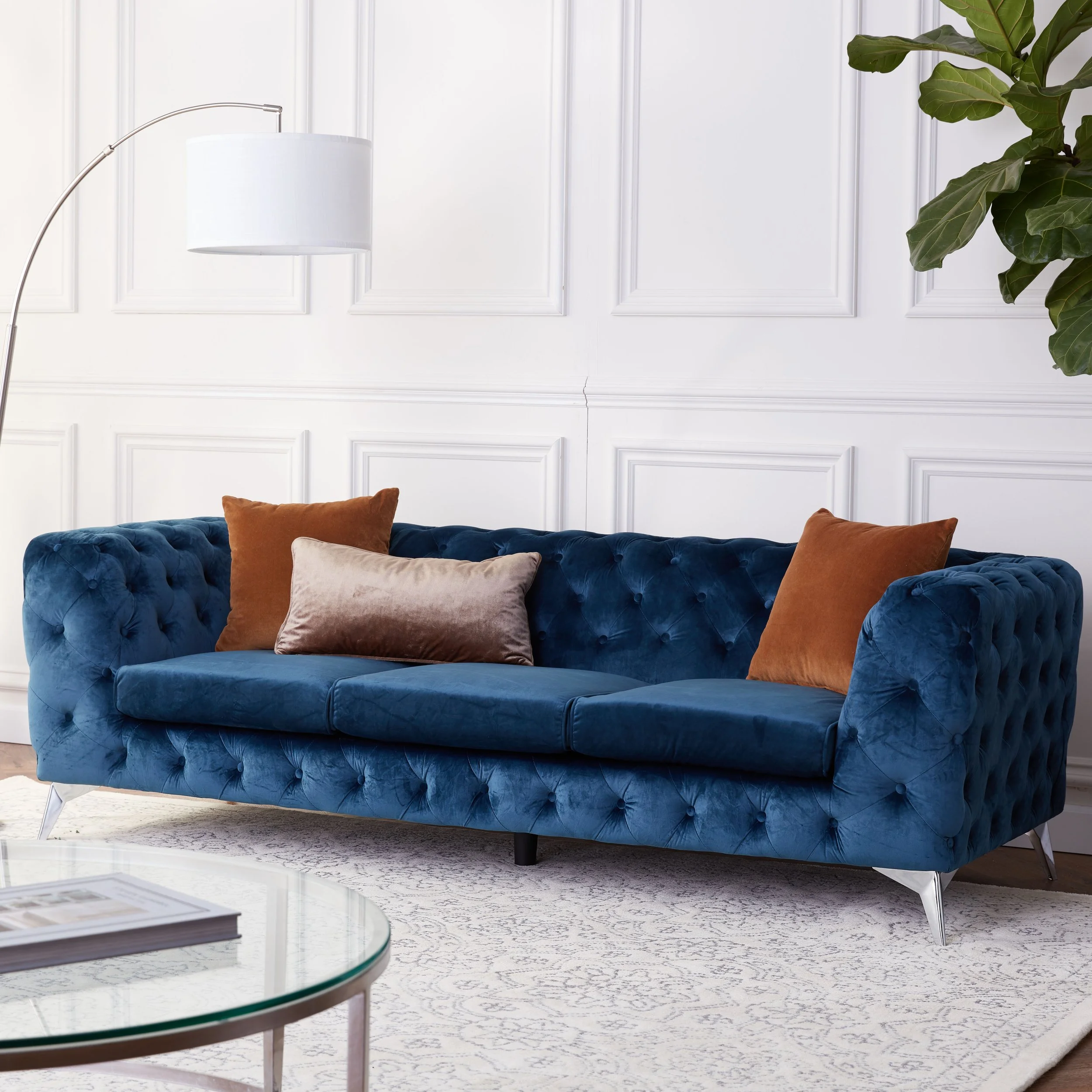 A blue velvet tufted sofa with three decorative pillows in front of a white paneled wall, with a large green plant partially visible on the right and a glass coffee table with magazines in the foreground.