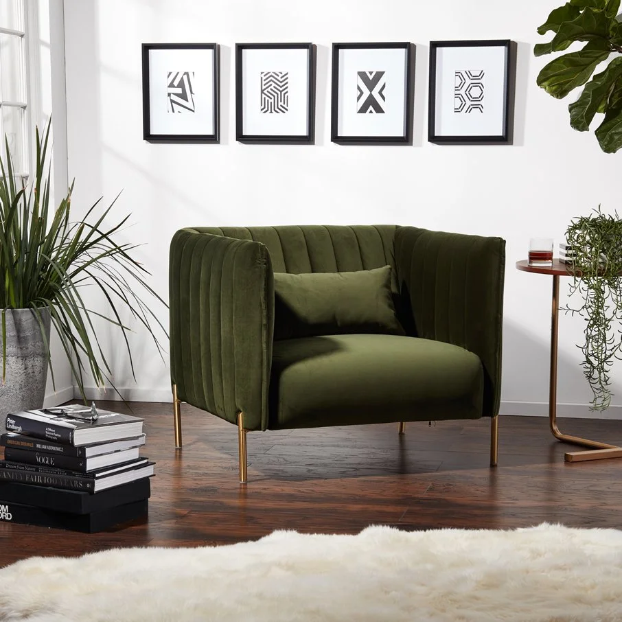 Living room with a green velvet armchair, framed abstract black and white art on the wall, potted plants, a side table with a glass of red drink, stack of books, and a white fluffy rug, bright natural lighting.