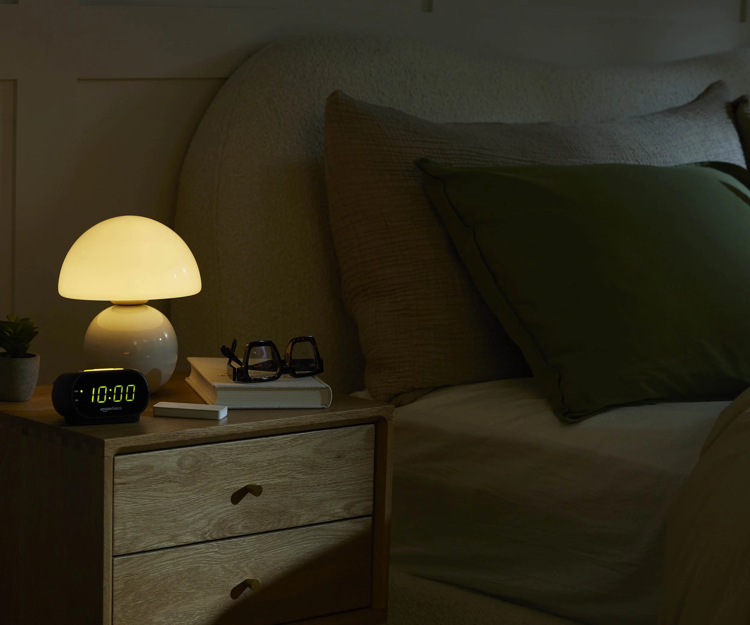 A cozy bedroom scene with a wooden nightstand, illuminated bedside lamp, a small digital alarm clock displaying 10:00, a pair of glasses, a notebook, and a stack of books. The bed has beige and green pillows, and the room has soft lighting.
