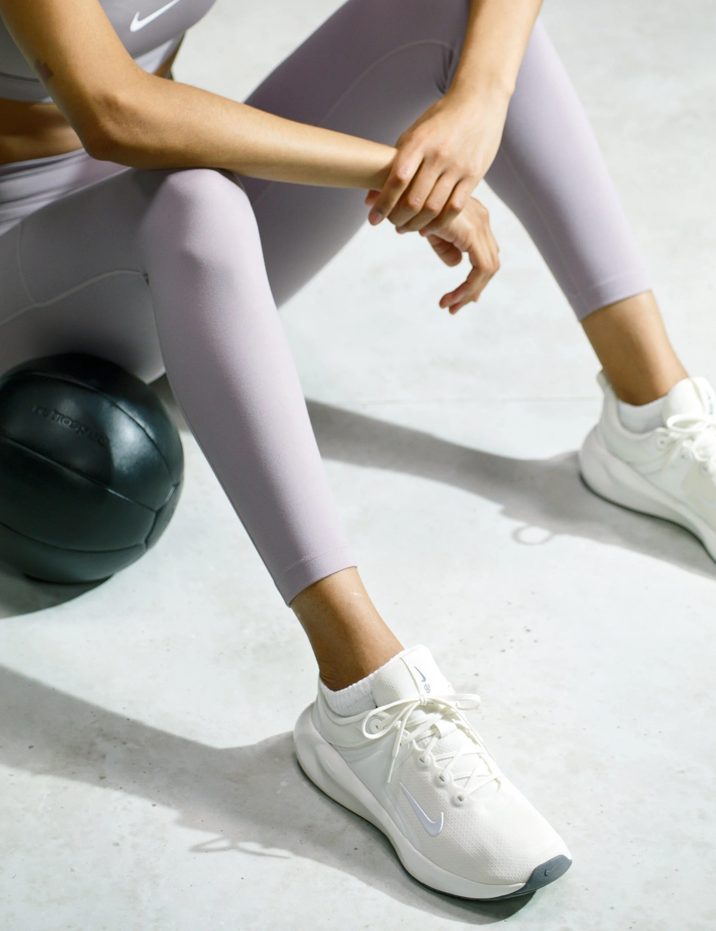 A person wearing white Nike sneakers, lavender leggings, and a gray athletic top sitting on a bench with a black medicine ball nearby, on a light-colored gym floor.