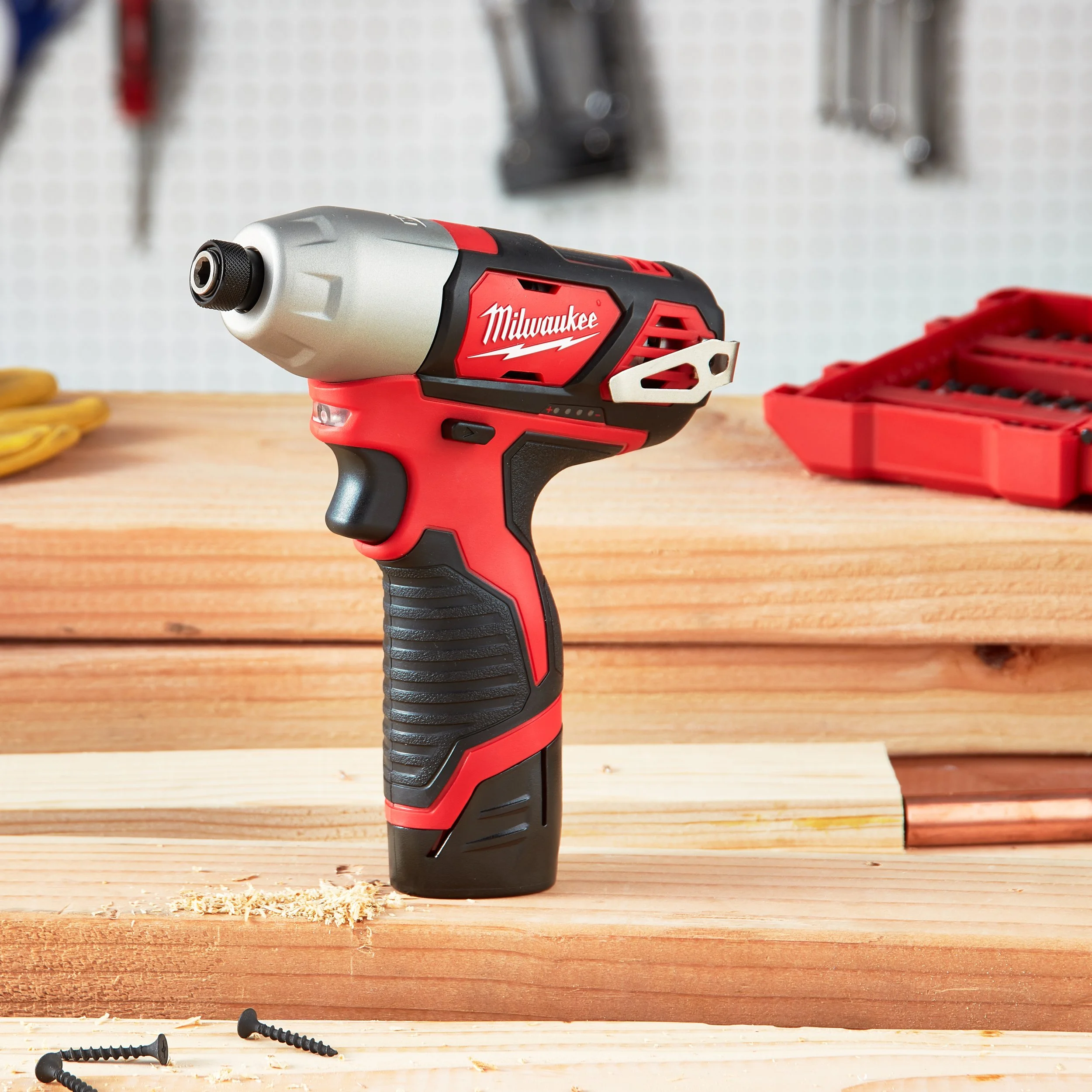 A red and black Milwaukee cordless power drill on a wooden work surface with screws and a red tool organizer in the background.