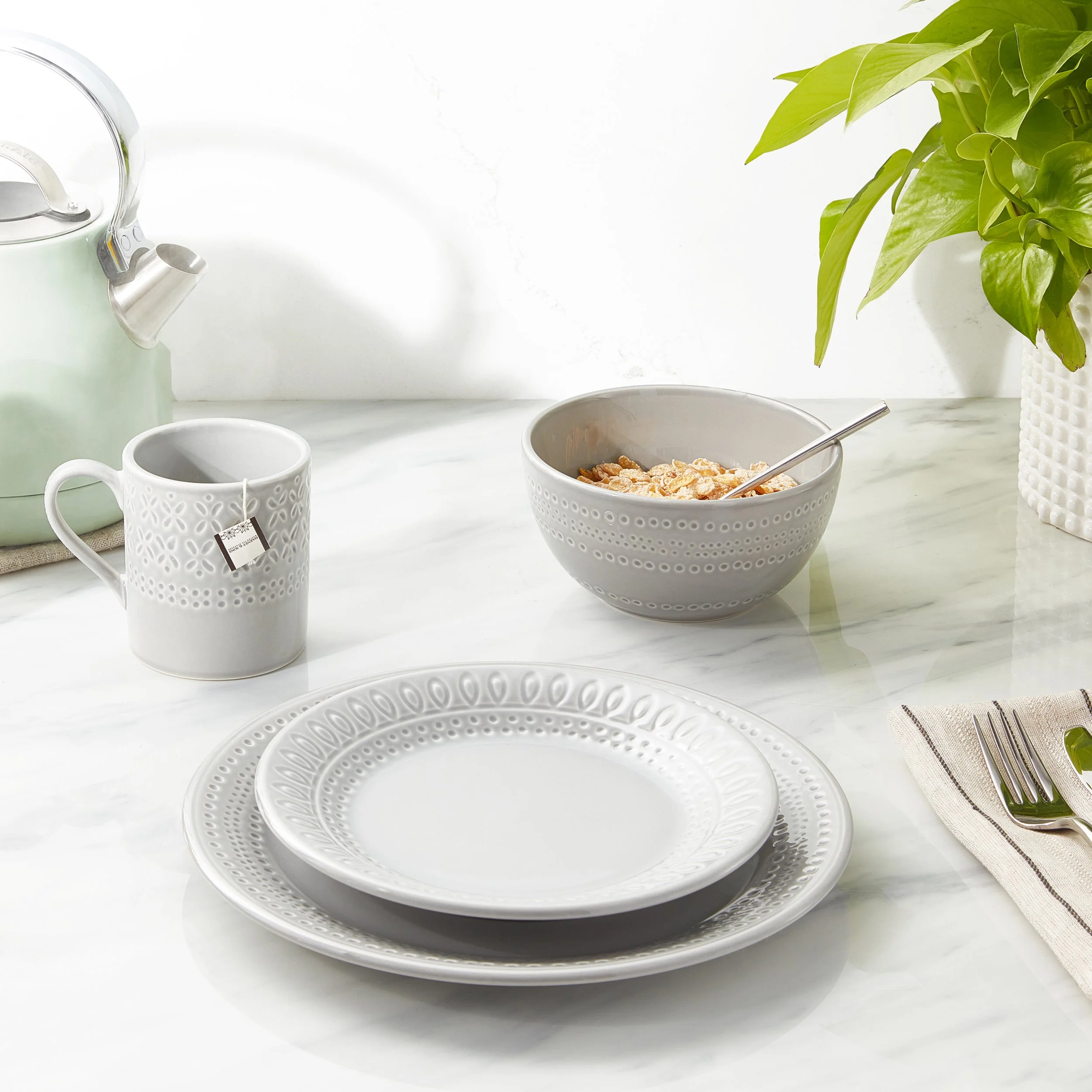 White ceramic plates, a bowl of cereal with milk and a spoon, a coffee mug, a teapot, and a plant on a marble table.